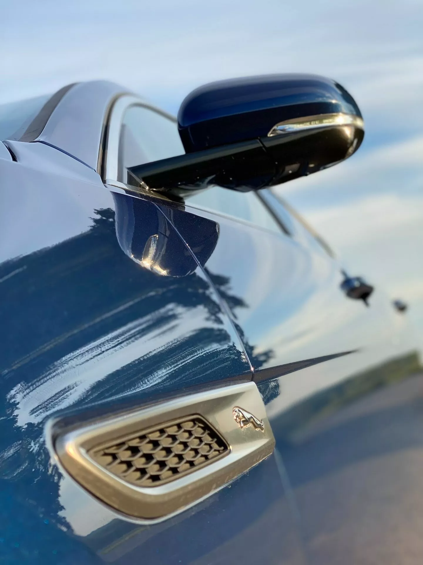 Zbliżenie na boczny błotnik i boczne lusterko Jaguara XF 2021 z niskiego kąta w górę, z głębokim metalicznym niebieskim lakierem odbijającym niebo. Wyraźne boczne lusterko, z błyszczącą czarną obudową, kontrastuje z kolorem nadwozia. Na błotniku widoczne jest logo producenta i kratka w kształcie plastra miodu, podkreślająca elementy designu i wydajności.