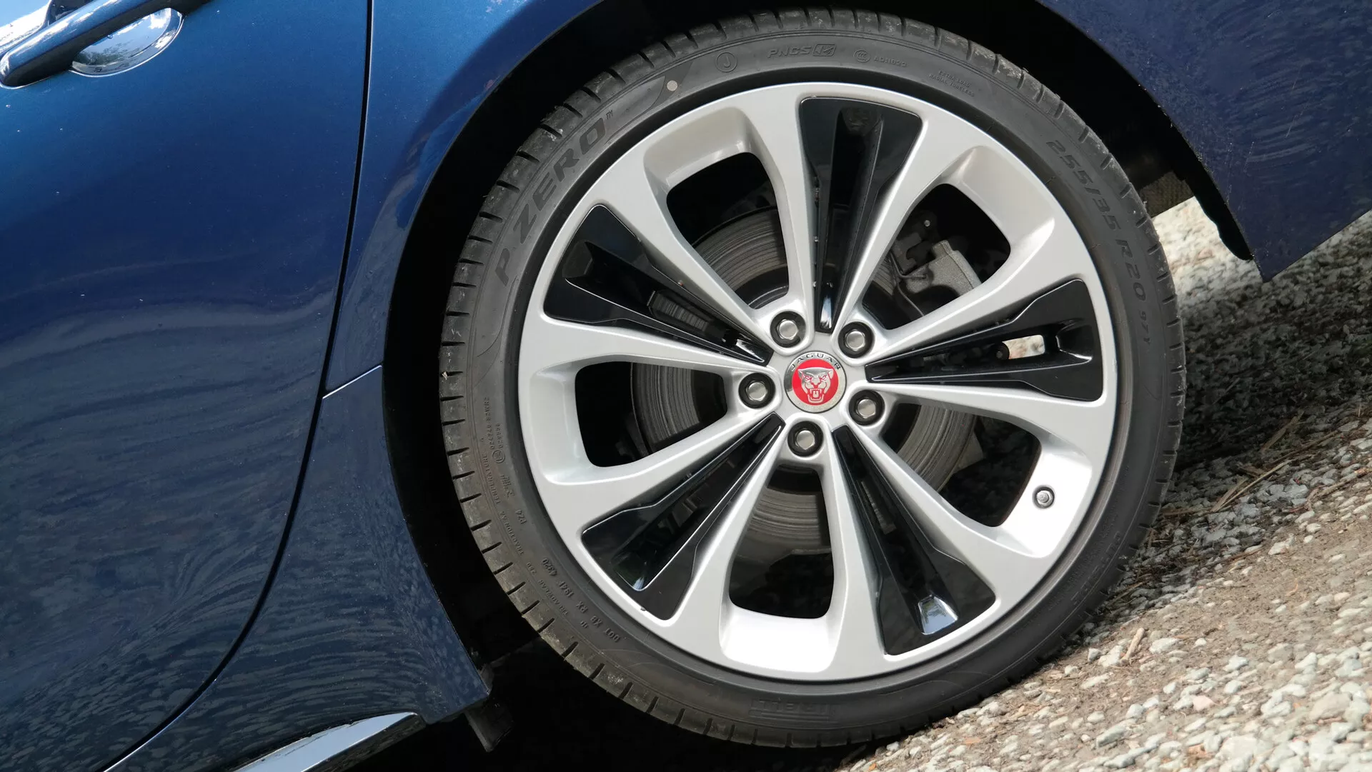 Zbliżenie na przednie koło po stronie kierowcy Jaguara XF 2021 z niskiego kąta, przedstawiające stylowy, wieloszprychowy design ze stopów metali z metalicznym srebrnym i ciemniejszym wykończeniem, szczegółowy wzór bieżnika opony oraz odblaskowe metaliczne wykończenie na dolnej części panelu bocznego samochodu.