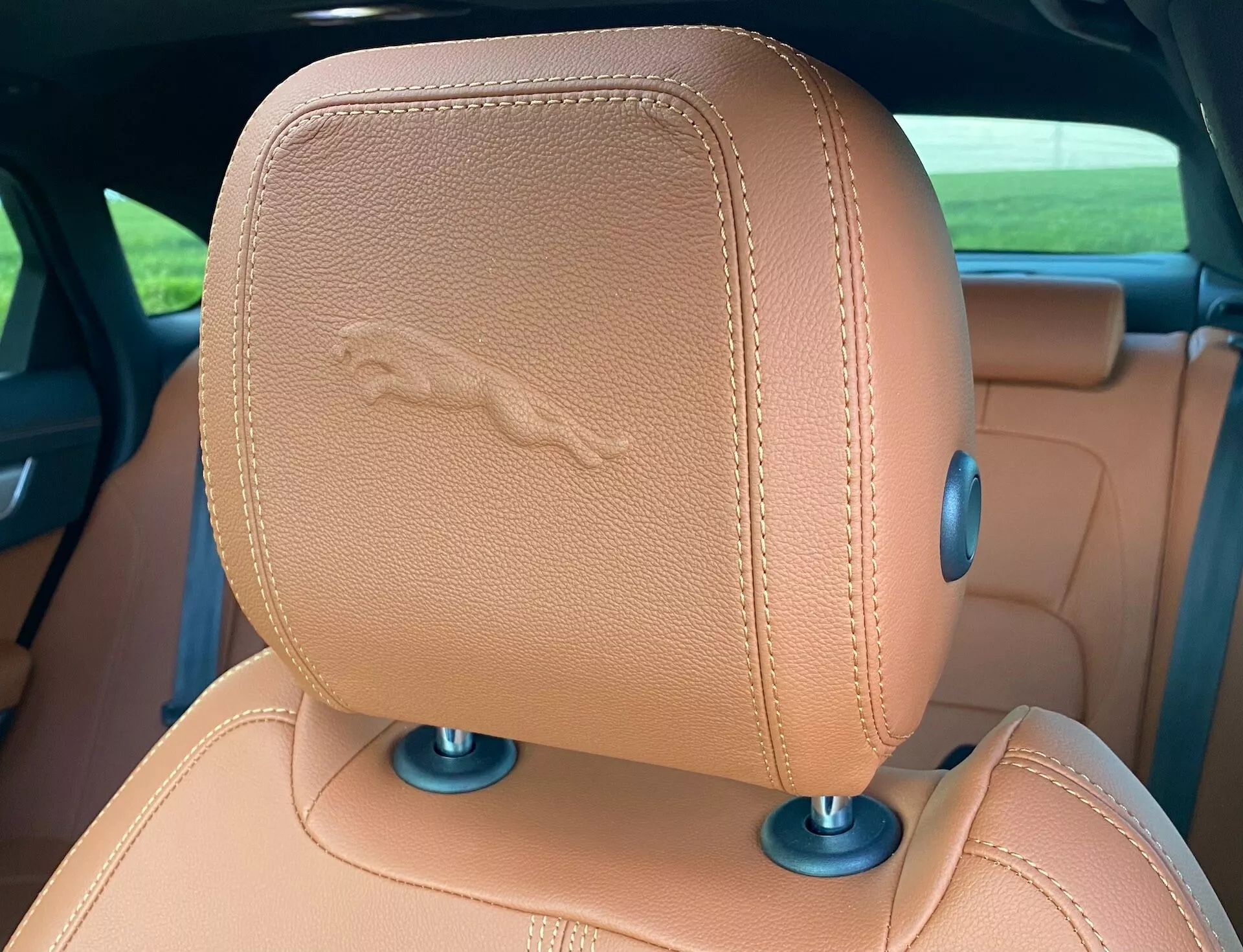 Zbliżenie na zagłówek oparcia fotela Jaguar XF 2021, z jasnobrązową skórą i wyszywanym emblematem konia, co sugeruje funkcje luksusowe. Niebieskie okrągłe elementy u podstawy wskazują na możliwe mechanizmy regulacyjne.