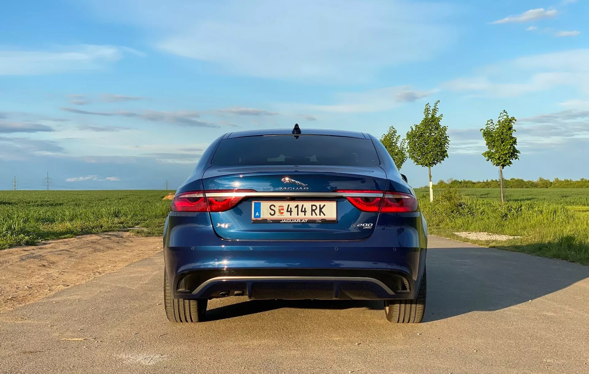 Tylny widok hatchbacka Jaguar XF 2021 na wąskiej asfaltowej drodze, światło dzienne z jasnym niebem, pola po obu stronach. Widoczne cechy: poziome tylne światła LED, podwójne wyloty wydechowe, centralne logo, emblemat modelu po prawej, centralna tablica rejestracyjna, mały tylny spojler, odblaski na tylnym zderzaku, możliwe czujniki parkowania/kamera, antena typu płetwa rekina.