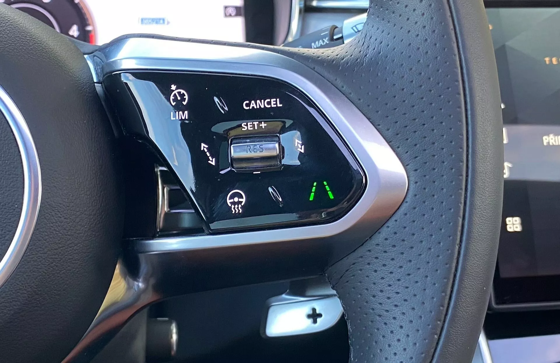 Zbliżenie na przyciski sterujące na kierownicy Jaguara XF 2021, pokazujące wielofunkcyjne przełączniki z perforowaną skórą dla lepszego uchwytu, metaliczne lub drewniane akcenty i różnorodne przyciski sterujące. Widoczne funkcje obejmują ustawienia wspomagania kierowcy dla ogranicznika prędkości i tempomatu z podświetleniem dla lepszej widoczności w warunkach słabego oświetlenia.