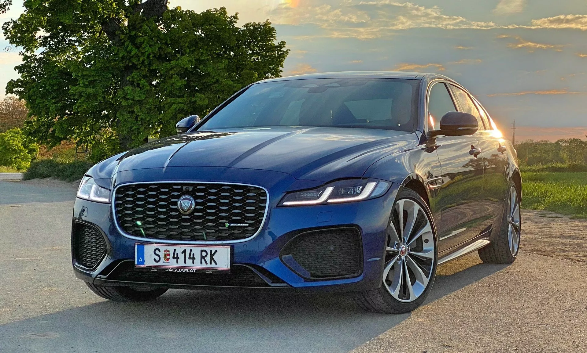 Nowoczesny sedan Jaguar XF 2021 w widoku trzy czwarte z przodu, ukazujący dużą przednią kratkę z emblematem producenta, eleganckie kątowe światła, sportowy przedni zderzak z wlotami powietrza i część bocznego profilu z podniesionymi liniami akcentującymi maskę, dużymi kołami ze stopu metali lekkich i lusterkami bocznymi z wbudowanymi wskaźnikami kierunku. Pojazd znajduje się na drodze z naturalnym krajobrazem w tle, na którym widać drzewa i wieczorne niebo podczas zachodu słońca.