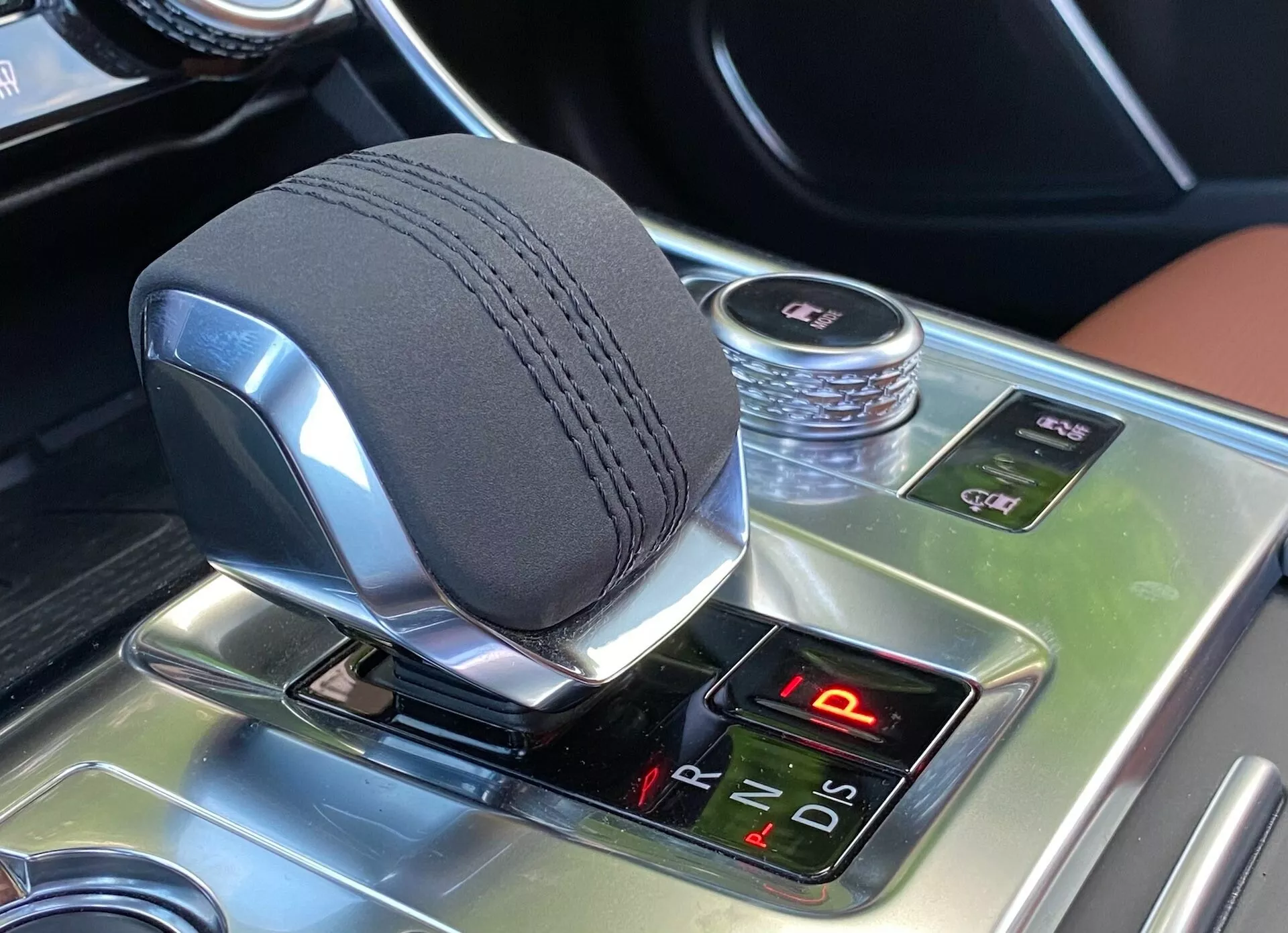 Zbliżenie wnętrza Jaguara XF 2021 z obszarem zmiany biegów, ukazujące elegancki, metaliczny elektroniczny selektor biegów shift-by-wire z podświetlanymi pozycjami biegów, otoczony błyszczącą konsolą środkową w efekcie drewna z zaawansowanymi funkcjami, w tym kontrolą trakcji, elektronicznym hamulcem postojowym oraz dezaktywacją systemu Auto Start/Stop.