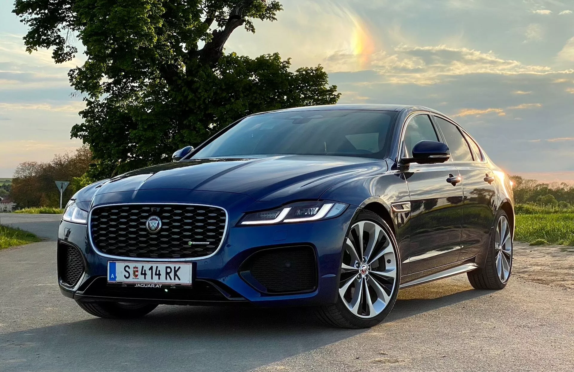 Sedan Jaguar XF 2021 uchwycony pod niskim przednim kątem trzech czwartych, podkreślający przednią fasadę, kratkę, maskę i profil strony kierowcy. Pojazd w głębokim niebieskim kolorze z wypolerowaną powierzchnią jest zaparkowany na asfaltowej drodze na tle wiejskiego krajobrazu z zachodzącym słońcem rzucającym ciepły blask. Wyraźna przednia kratka ma charakterystyczny wzór, otoczona eleganckimi reflektorami LED. Przedni zderzak prezentuje wloty powietrza i możliwe czujniki systemów wspomagania kierowcy, podkreślone niskim spojlerem. Wieloramienne felgi aluminiowe z oponami o niskim profilu podkreślają zaprojektowanie pojazdu zorientowanego na osiągi.