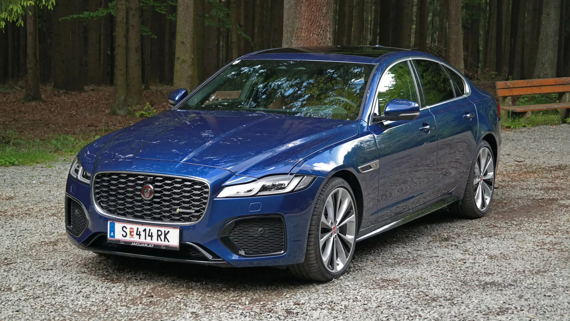 Obraz przedstawia Jaguara XF 2021, nowoczesnego luksusowego sedana zaparkowanego na żwirowej nawierzchni w lesistej okolicy. Uchwycony pod kątem trzech czwartych od przodu, zdjęcie ukazuje wyraźny przedni grill samochodu z centralnym emblematem, otoczony eleganckimi reflektorami LED, prawdopodobnie z wbudowanymi światłami do jazdy dziennej i być może technologią adaptacyjną. Poniżej reflektorów widać duże wloty powietrza, które mogą spełniać zarówno funkcje estetyczne, jak i praktyczne. Maskę charakteryzuje rzeźbiony design z wypukłymi krawędziami, a pochylona szyba sugeruje aerodynamiczną efektywność. Profil boczny ujawnia duże aluminiowe felgi z wieloszprychowym designem i niskoprofilowe opony, co może wskazywać na osiągi. Linia karoserii biegnie wzdłuż boku, dodając wrażenia ruchu, podczas gdy lusterka boczne w kolorze nadwozia z wbudowanymi kierunkowskazami oraz okna z chromowanymi obramowaniami podkreślają luksusowy wygląd pojazdu.