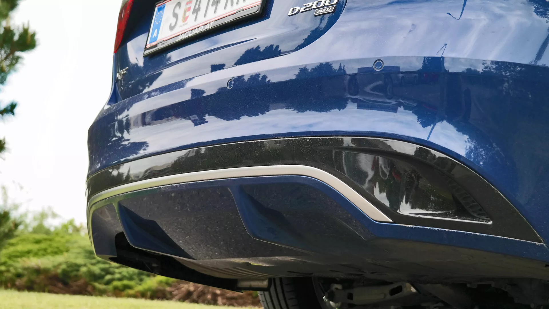 Zbliżenie tylnej części niebieskiego Jaguara XF 2021 uchwyconego pod ukośnym kątem z poziomu gruntu, ukazującego zintegrowany czarno-srebrny dyfuzoryczny zderzak, metaliczny niebieski lakier z otaczającymi odbiciami oraz odznakę 'DDDI' na lewym boku pokrywy bagażnika, bez widocznych świateł tylnych ani rur wydechowych.