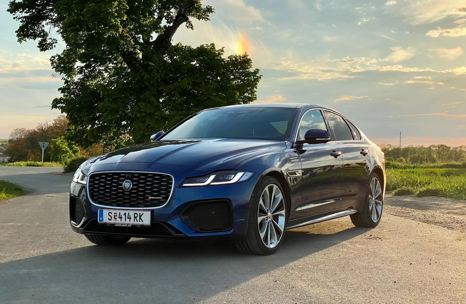 Ten obraz przedstawia luksusowego sedana Jaguar XF 2021, ustawionego pod kątem na wiejskiej drodze. Samochód jest skierowany lekko w prawo kadru, ukazując jego przedni lewy ćwiartka, w tym dużą kratkę z siatką, eleganckie reflektory LED, wlot powietrza zintegrowany z czujnikami parkowania, lusterka boczne montowane na drzwiach prawdopodobnie ze wskaźnikami kierunku, wieloszprychowe felgi aluminiowe z oponami o niskim profilu i aerodynamiczne kontury karoserii z eleganckim profilem. Scena osadzona jest w spokojnym, wiejskim otoczeniu z ciepłym, naturalnym wieczornym oświetleniem, podkreślając premiowy design i zaawansowane technologicznie funkcje samochodu.