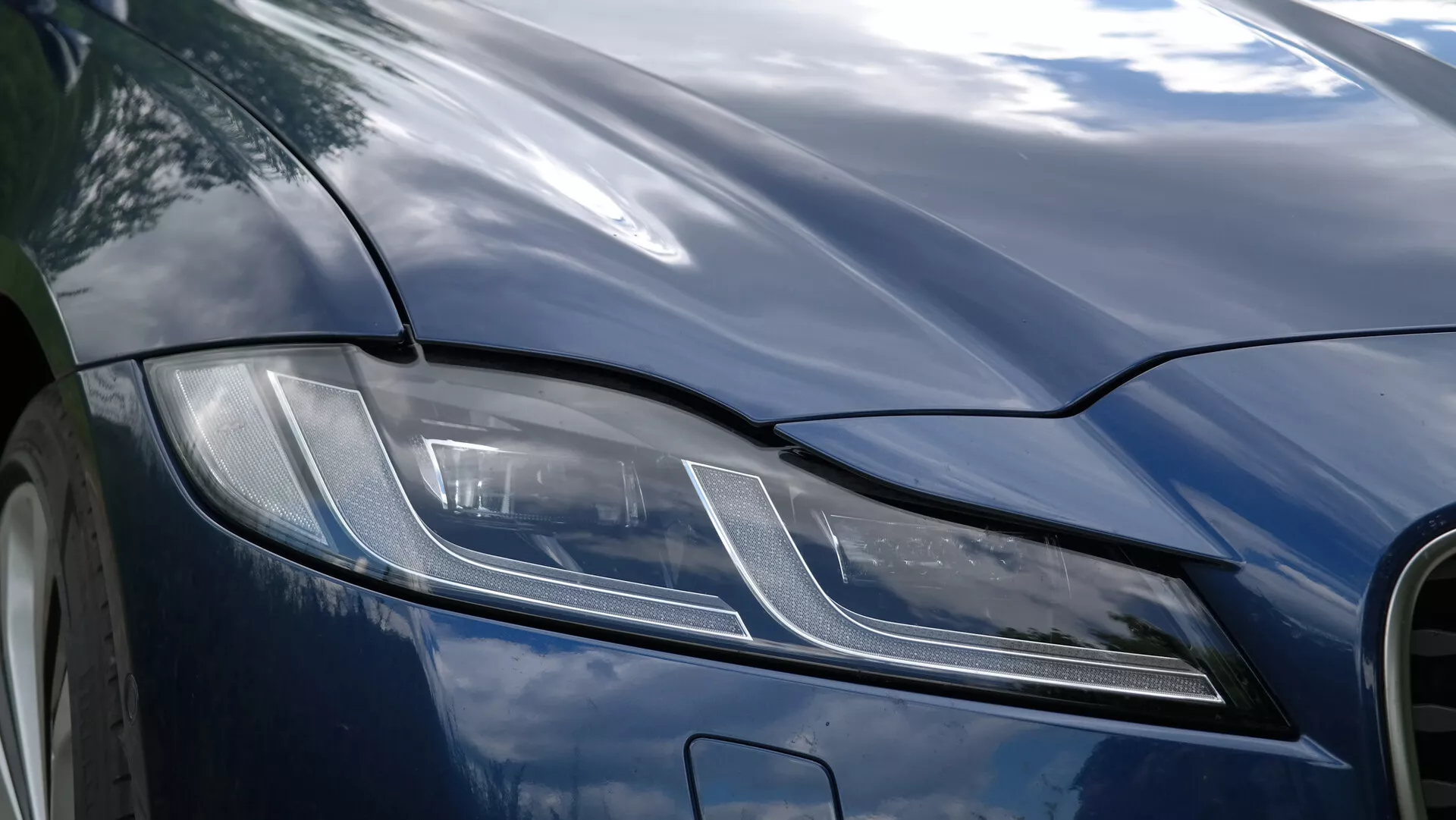 Zbliżenie na przedni lewy kwartał Jaguara XF 2021, skupiające się na kątowych reflektorach LED, charakteryzujących się nowoczesnym wzornictwem z poziomymi paskami i kropkami, polerowaną ciemną maską i aerodynamicznymi liniami, obrazującymi luksus i zaawansowaną technologię motoryzacyjną.