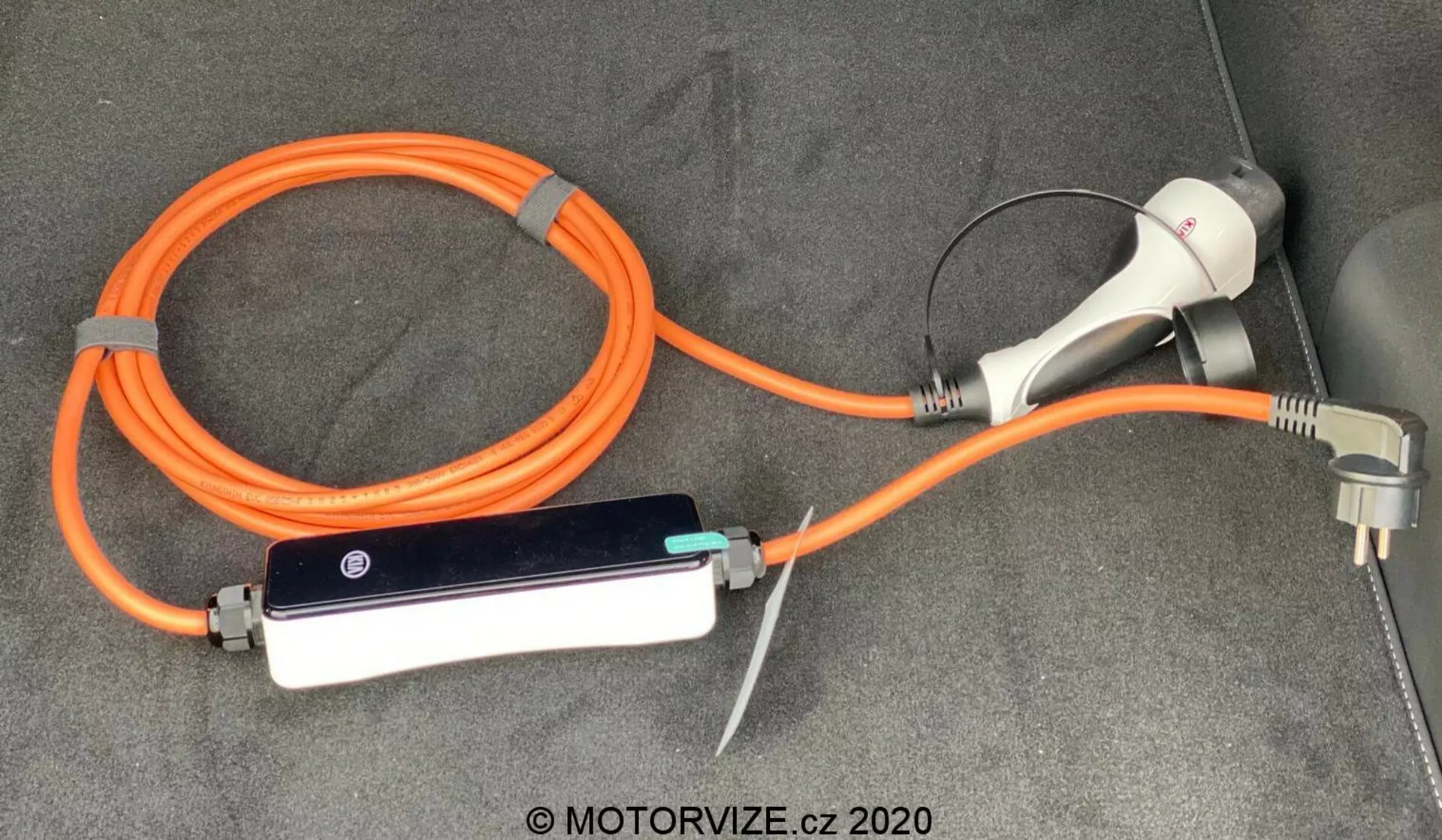 Zestaw kabli i adapterów do ładowania pojazdu elektrycznego (EV) dla Kia Ceed 2018, obejmujący przenośne, pomarańczowe EVSE (Urządzenie Zaopatrzenia w Energii Pojazdu Elektrycznego) o wysokiej widoczności dla bezpieczeństwa. Zestaw zawiera złącze i wtyczkę do ładowania do użytku domowego oraz kontroler z wskaźnikami LED, umieszczone na wykładzinie w bagażniku lub przestrzeni ładunkowej pojazdu.