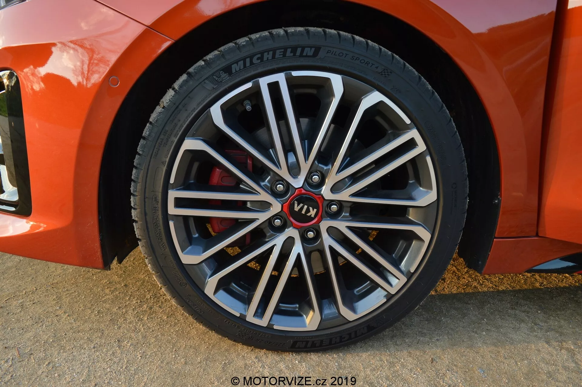 Zbliżenie przedniego koła pasażera Kia Ceed 2021, pod lekkim kątem ukośnym, przedstawiające wieloramienną felgę aluminiową z ciemniejszymi wewnętrznymi sekcjami i jasnymi maszynowymi krawędziami. Posiada oznakowany piast z czerwonym znakiem 'VT1', oponę Michelin z szczegółowymi informacjami na ścianie boczne oraz czerwone zaciski hamulcowe wskazujące na komponenty o wysokiej wydajności. Miękkie cienie i oświetlenie sugerują, że zdjęcie zostało wykonane w naturalnym świetle, prawdopodobnie podczas złotej godziny.