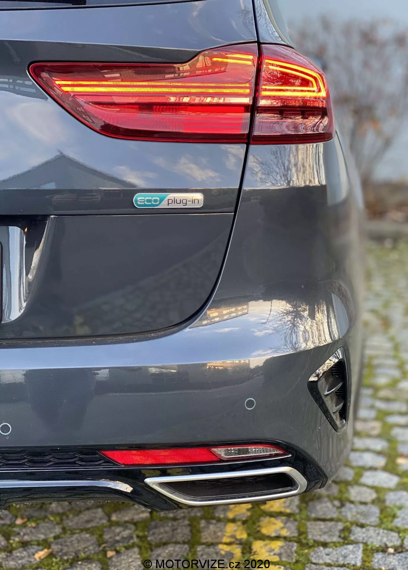 Szczegółowy widok tylnej części Kia Ceed 2018 w metalicznym szarym kolorze, podkreślający podświetlane tylne światło LED, emblemat eco plug-in, rzeźbiony tylny zderzak z reflektorem i dyfuzorem oraz podwójne chromowane końcówki wydechu. Perspektywa pokazuje zbieżność tylnego zderzaka i prawej strony, podkreślając elementy hybrydowego pojazdu elektrycznego i nowoczesne cechy stylistyczne.