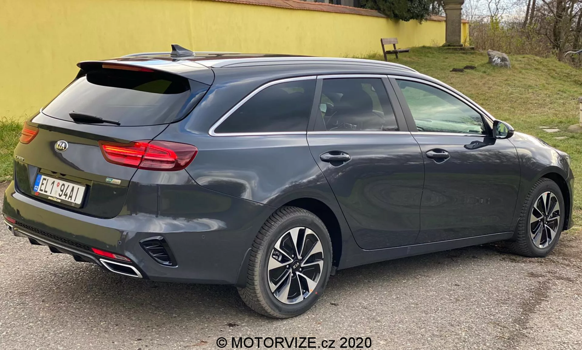 Ciemnoszary metaliczny Kia Ceed 2018, pokazany z trzech czwartych tyłu, podkreślając nowoczesny design hatchback lub kombi z pochyłym tylnym oknem, zintegrowanym tylnym spojlerem, wieloramiennymi felgami aluminiowymi, klamkami i lusterkami bocznymi w kolorze nadwozia prawdopodobnie z wbudowanymi kierunkowskazami, charakterystyczne poziome tylne światło prawdopodobnie z technologią LED, anteną w kształcie płetwy rekina, podwójnymi rurami wydechowymi, wysoko zamontowaną trzecią lampą stopu, funkją odmrażania tylnej szyby, czujnikami lub odblaskami wskazującymi na system wspomagania parkowania z tyłu, fotografowany z bliska w neutralnym oświetleniu.