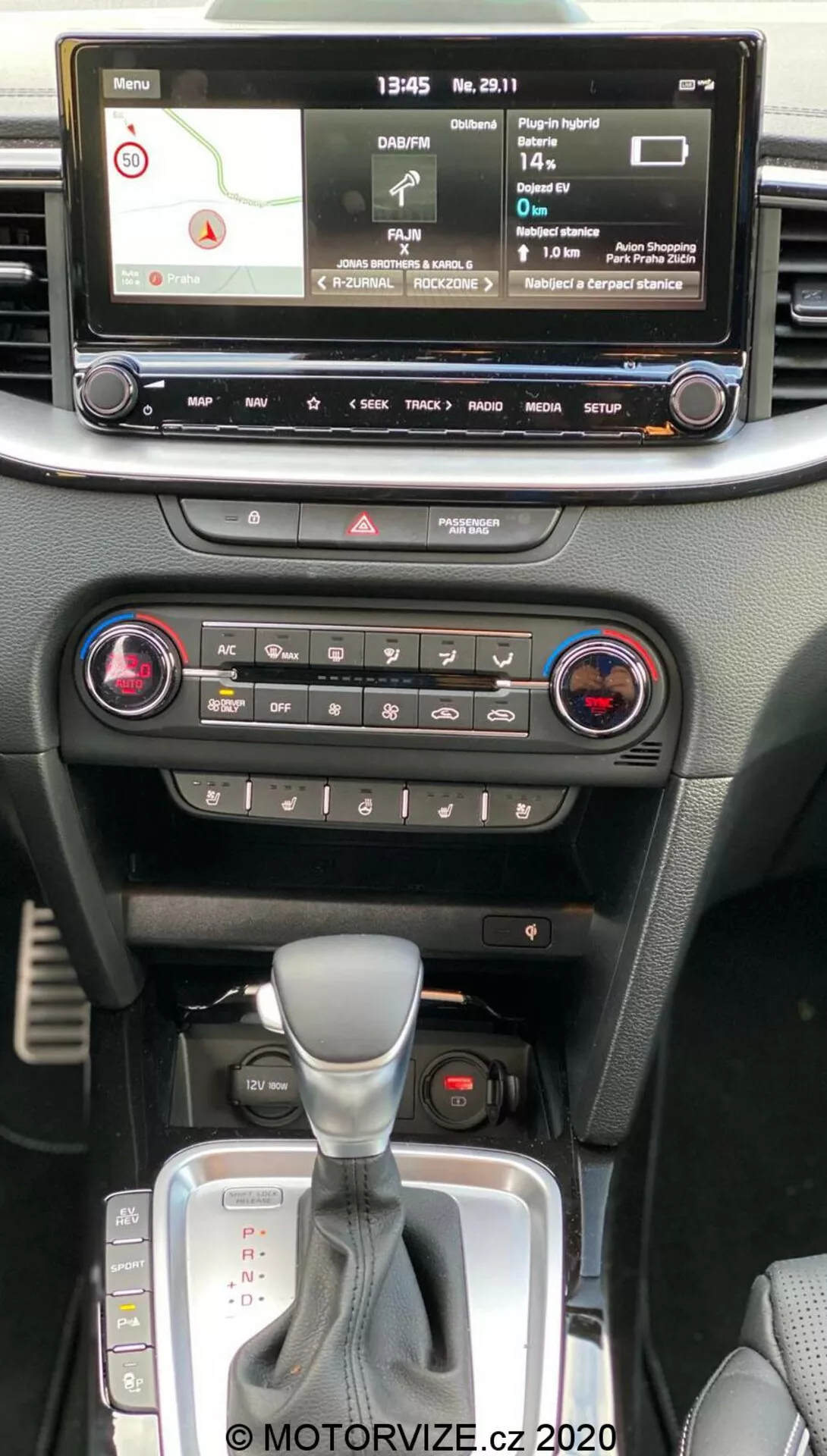 Wnętrze Kia Ceed 2018 pokazujące konsolę centralną z perspektywy pasażera w pierwszym rzędzie, z cyfrowym wyświetlaczem infotainment z mapą nawigacji, odtwarzaczem multimediów, ustawieniami pojazdu, sterowaniem klimatyzacją z systemem dwustrefowym, przyciskiem świateł awaryjnych, automatyczną skrzynią biegów z osłoną skórzaną i slotem na kluczyk do uruchamiania bezkluczykowego.