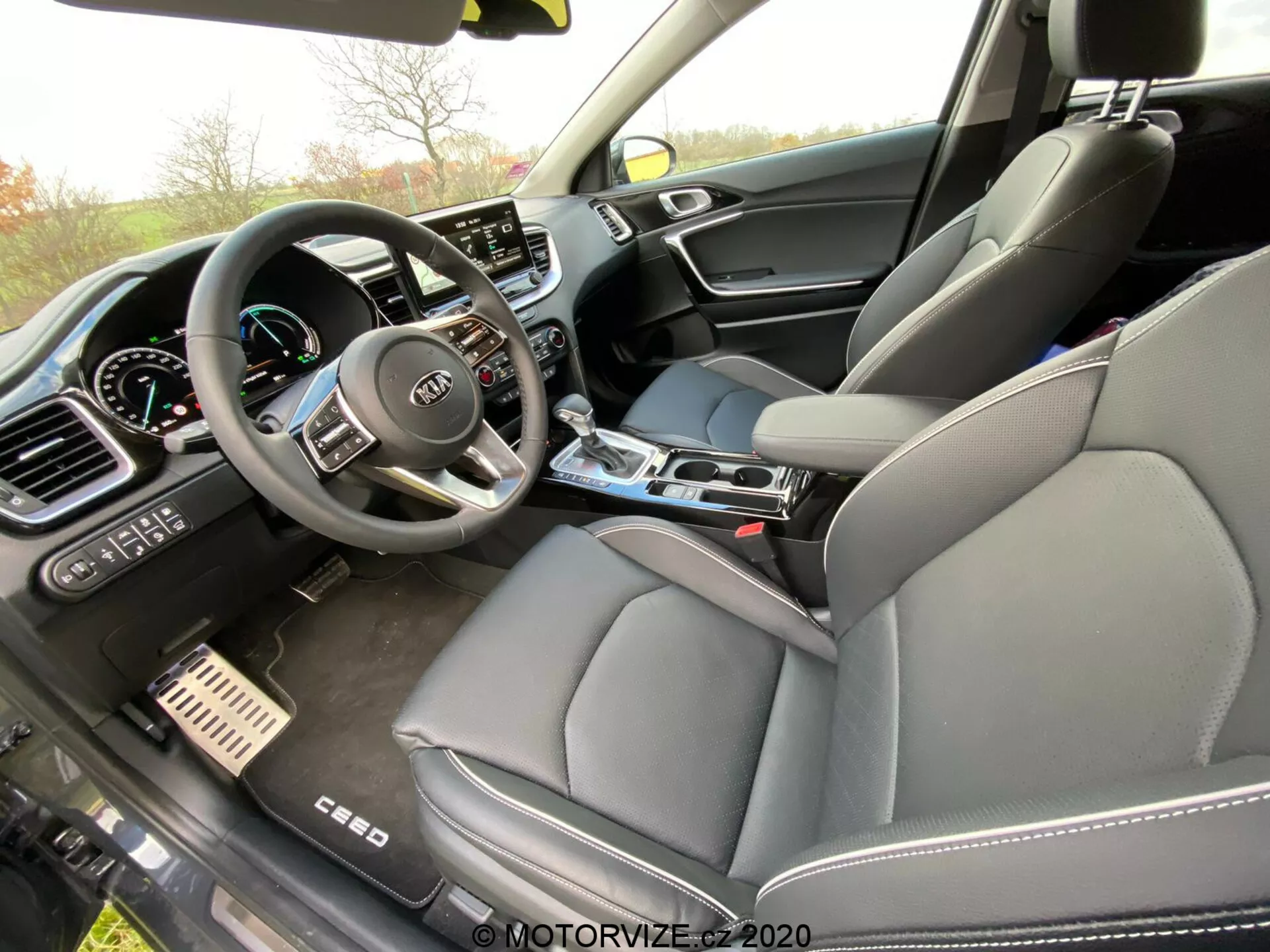 Zdjęcie wnętrza Kia Ceed 2018 z perspektywy pasażera, uchwyte przednią część z naciskiem na deskę rozdzielczą, konsolę środkową i fotel kierowcy. Widoczna jest kierownica z wieloma przyciskami sterującymi i logo Kia, wraz z częściowo widocznym cyfrowym zestawem wskaźników. Środkowa część deski rozdzielczej zawiera duży ekran dotykowy systemu informacyjno-rozrywkowego otoczony manualnymi ustawieniami klimatyzacji. Konsola środkowa zawiera dźwignię zmiany biegów, uchwyty na kubki i pokrętła sterowania. Widać również przedni fotel pasażera obity skórą i mieszany materiał na panelu drzwi, co sugeruje luksusowe wykończenie. Metalowe pedały hamulca i przyspieszenia również wskazują na sportowy, premium styl.