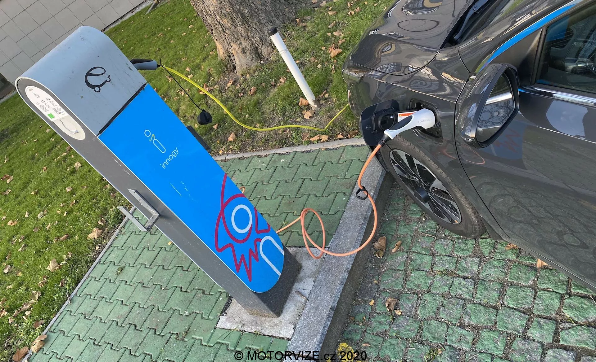 Widok z góry na nowoczesny pojazd elektryczny ładowany przy publicznej stacji ładowania. Obraz ukazuje prawą tylną część samochodu, skupiając się na porcie ładowania z podłączonym grubym pomarańczowym kablem. Stacja ładowania ma niebieską fasadę z grafiką iskry elektrycznej i wyświetlaczem u góry, sugerującym elementy interfejsu użytkownika. Scena jest osadzona w zielonym otoczeniu przypominającym park, wskazując na wygodne miejskie lub podmiejskie położenie.
