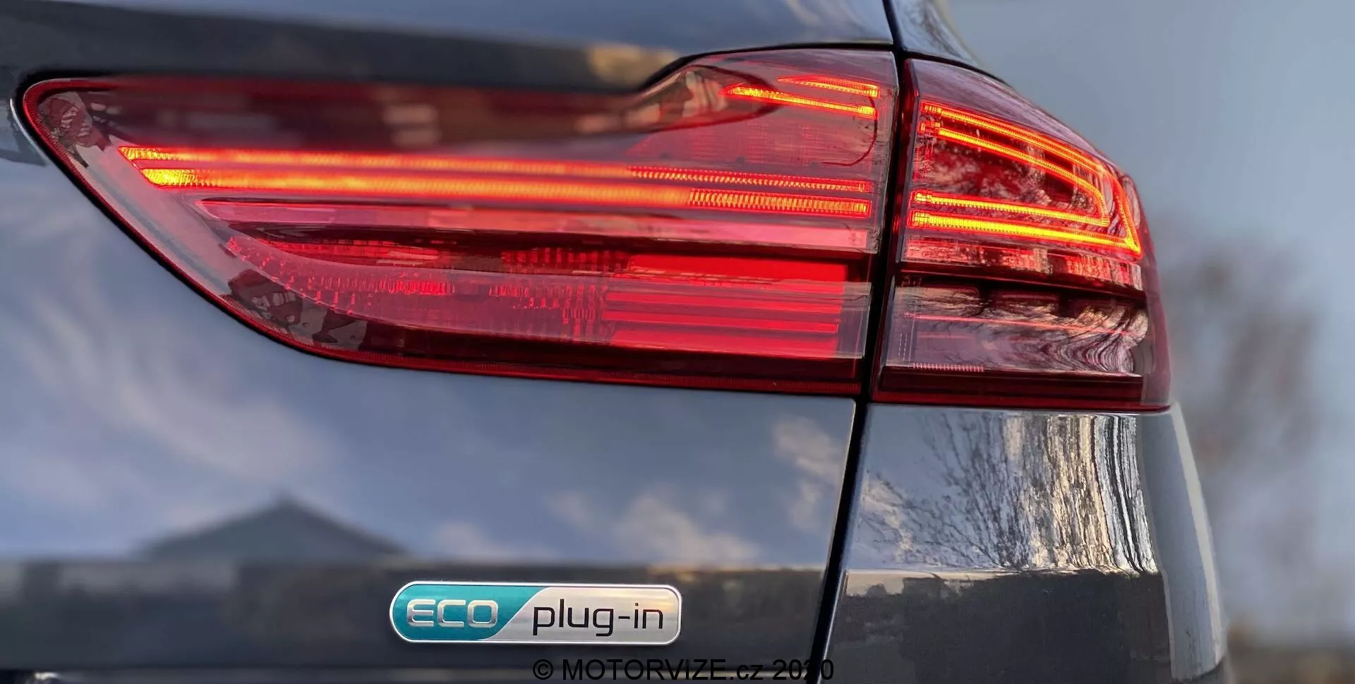 Zbliżenie tylnej lewej strony Kia Ceed 2018, skupiające się na podświetlonych tylnych światłach z wielowarstwowym wzornictwem. Odznaka 'ECO plug-in' wskazuje, że jest to pojazd hybrydowy typu plug-in. Błyszczące wykończenie i lekko widoczna klapa bagażnika dodają szczegółów do ogólnego designu samochodu.