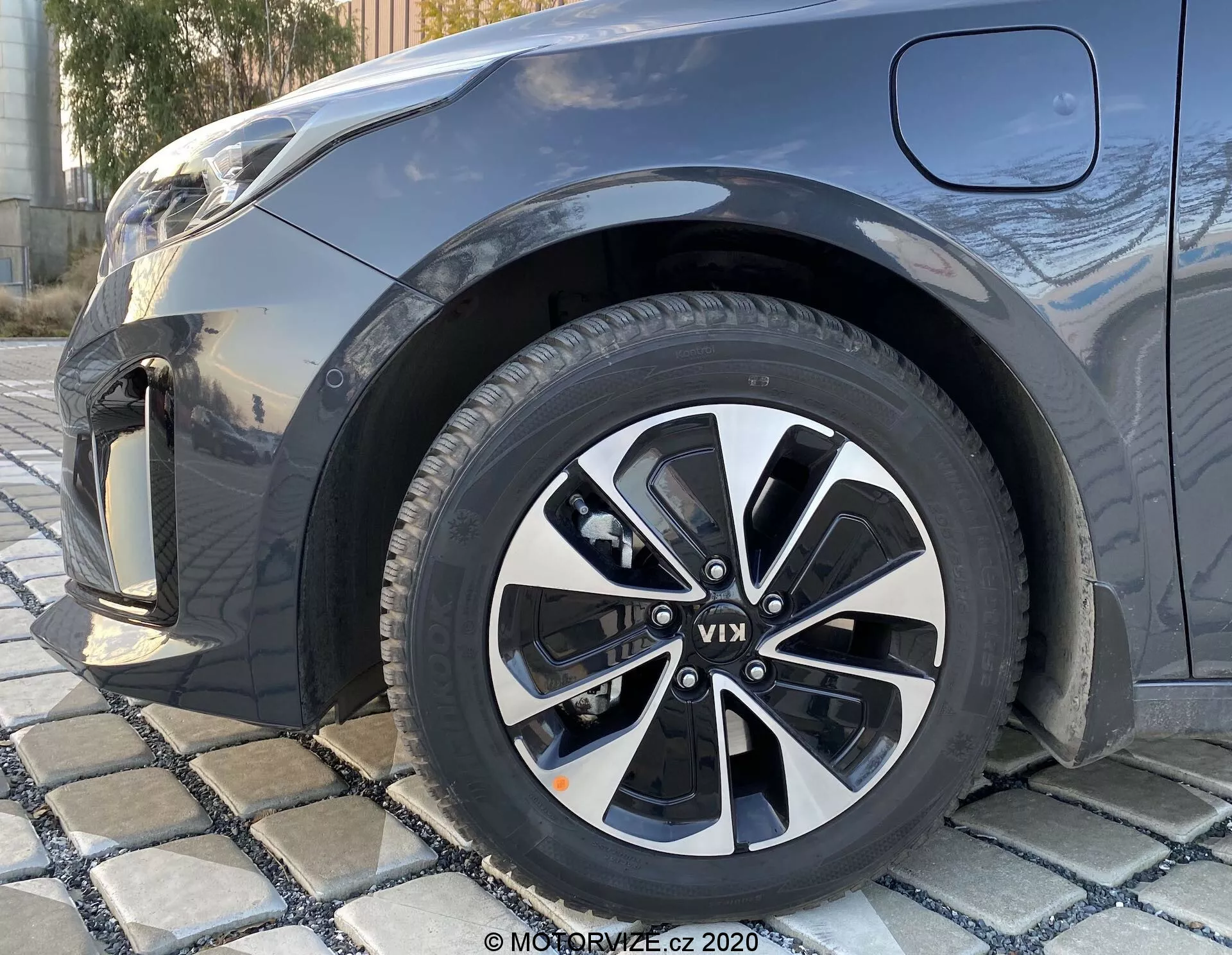 Zbliżenie przedniego prawego koła Kia Ceed 2018 z niskiego kąta, z wieloramiennym wzorem felg w kolorze czarnym i srebrnym, z widocznymi wzorami bieżnika, częściowo ukazując przedni błotnik, zderzak i część maski w ciemnym metalicznym kolorze.