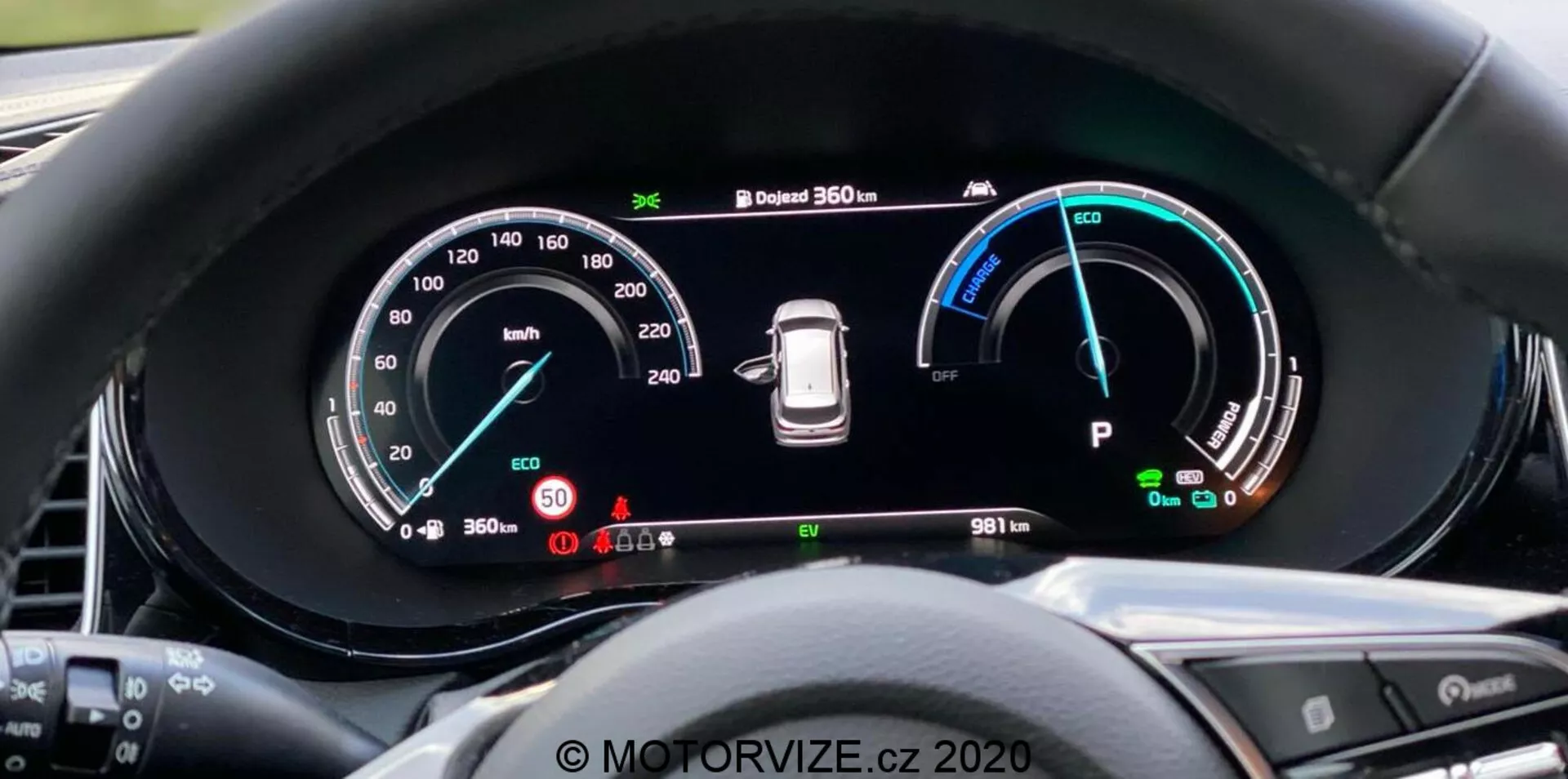 Zbliżenie na cyfrową tablicę rozdzielczą Kia Ceed 2018 z perspektywy kierowcy, z dużym panoramicznym ekranem i szczegółową instrumentacją, w tym prędkościomierzem do 260 km/h, wskaźnikiem mocy z trybami Eco, Comfort i Power, ostrzeżeniem o ciśnieniu w oponach i wskaźnikiem trybu 'Park'. Tablica rozdzielcza pokazuje również system kamer 360 stopni i licznik kilometrów z odczytem 9811 km. Otaczająca krawędź zmniejsza odblaski, a przyciski na kierownicy są częściowo widoczne. Znak wodny 'MOTORVIZE.cz 2020'.