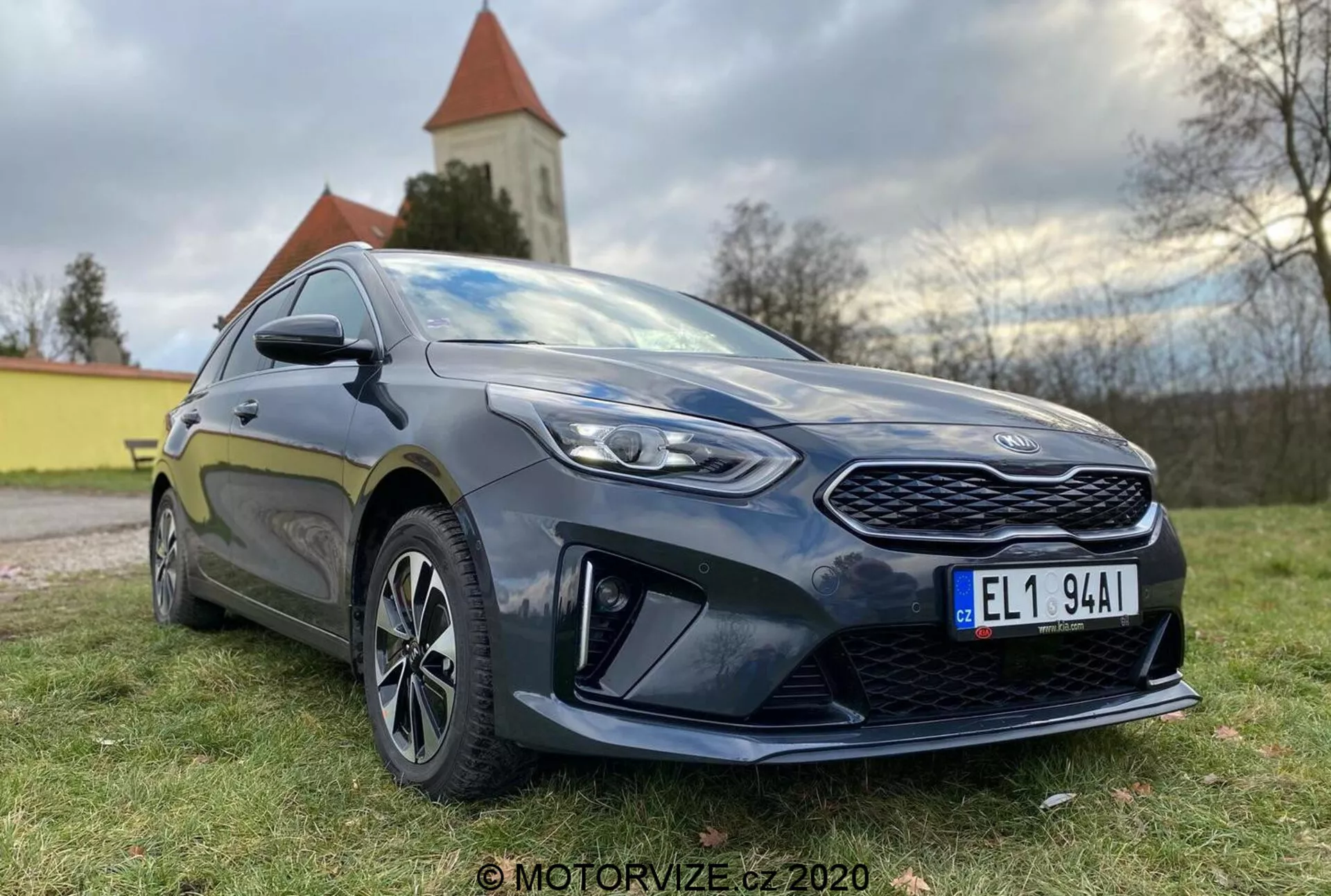 Szczegółowy widok nowoczesnego hatchbacka Kia Ceed 2018, sfotografowanego pod niskim kątem przednim trzy czwarte, ukazującego wyraźny grill, kanciaste reflektory LED i boczny profil do drzwi kierowcy. Samochód jest ciemny, prawdopodobnie ciemnoniebieski lub czarny, z połyskiem podkreślającym jego dobrze utrzymany stan. Charakterystyczne cechy projektu obejmują wzorzystą przednią kratę, ostre reflektory z przezroczystymi soczewkami, wyraźne elementy aerodynamiczne na dolnym przednim zderzaku, wieloramienne felgi aluminiowe, boczne lusterka w kolorze nadwozia z wbudowanymi kierunkowskazami i sportowy próg boczny. Postawa samochodu sugeruje równowagę między komfortem a sportowym charakterem. Naturalne światło dzienne z miękkim oświetleniem podkreśla detale, a w tle widoczny jest naturalny krajobraz z kościołem lub wieżą.