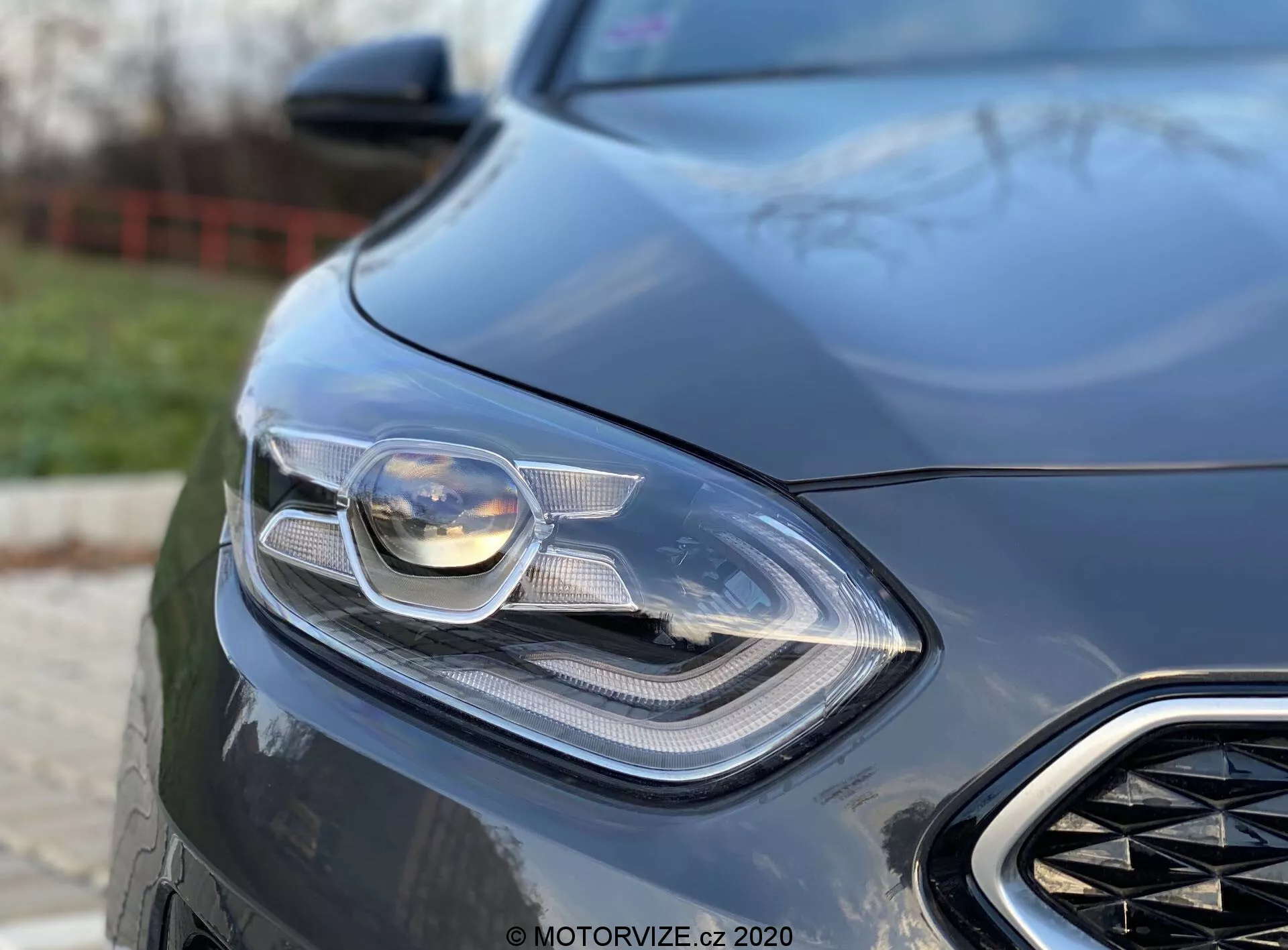 Szczegółowe zbliżenie na przednią lewą część Kia Ceed 2018, skupiając się na wyrafinowanym reflektorze i części przedniego grilla. Zdjęcie zrobione pod niskim kątem, podkreślające elegancki i nowoczesny design samochodu z widoczną zaawansowaną technologią oświetleniową, prawdopodobnie w tym reflektory LED i adaptacyjne. Rozmyte tło podkreśla aerodynamiczne elementy przedniego auta i luksusowy wygląd, wskazując na pojazd nastawiony na osiągi.
