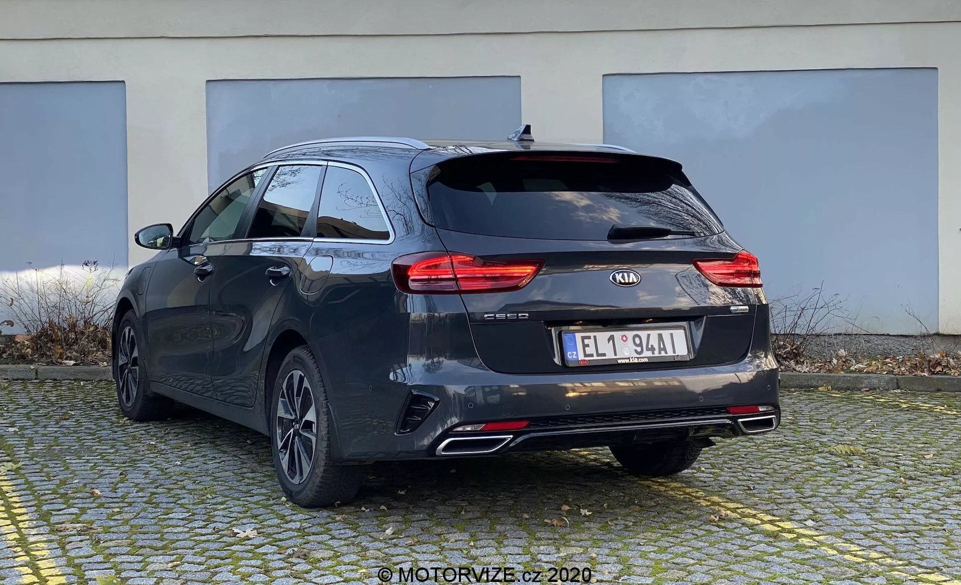 Obraz przedstawia Kia Ceed 2018 z widoku tylno-bocznego, ukazujący tył i bok kombi. Samochód ma ciemny kolor, prawdopodobnie czarny lub ciemnoszary, z charakterystycznymi liniami i lekko opadającym dachem, nadającym sportowy wygląd. Widać nowoczesne tylne światła LED, logo Kia, nazwę modelu 'CEED', dodatkowe oznaczenia, europejską tablicę rejestracyjną, tylne koła z wieloszprychowymi felgami, podwójne rury wydechowe i tylne wycieraczki. Sugerowane, ale niewidoczne są kamera cofania i czujniki parkowania. Mały spojler dachowy i relingi wskazują na dodatkowe opcje ładunkowe. Samochód jest zaparkowany na wybrukowanym obszarze z budynkiem w tle, pokazując jego tylny i częściowo boczny profil z lekkiego niskiego kąta.