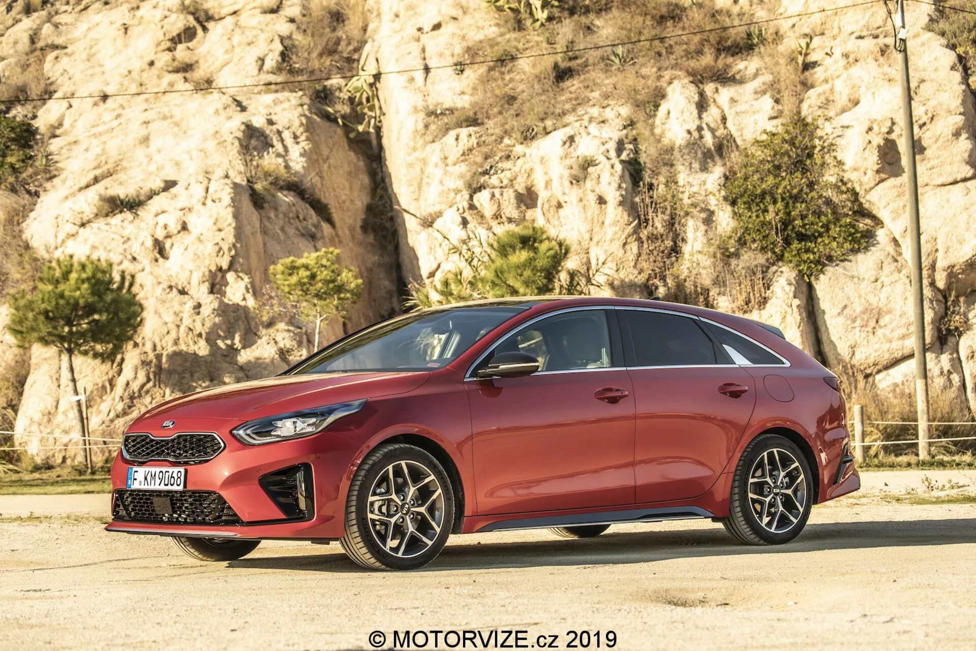 Widok z profilu bocznego czerwonego hatchbacka Kia Ceed 2021, z eleganckim, aerodynamicznym designem, opadającymi liniami dachu i konturowanymi liniami na drzwiach. Samochód, model pięciodrzwiowy, ma chromowane wykończenie okien i czarne słupki B, tworzące efekt ciągłej tafli szkła. Wieloszprychowe felgi aluminiowe z widocznymi tarczami hamulcowymi dodają sportowego charakteru. Ostro zarysowane reflektory i częściowo widoczna atrapa chłodnicy zaznaczają przód, a lusterka boczne w kolorze nadwozia z wbudowanymi kierunkowskazami poprawiają widoczność i bezpieczeństwo.