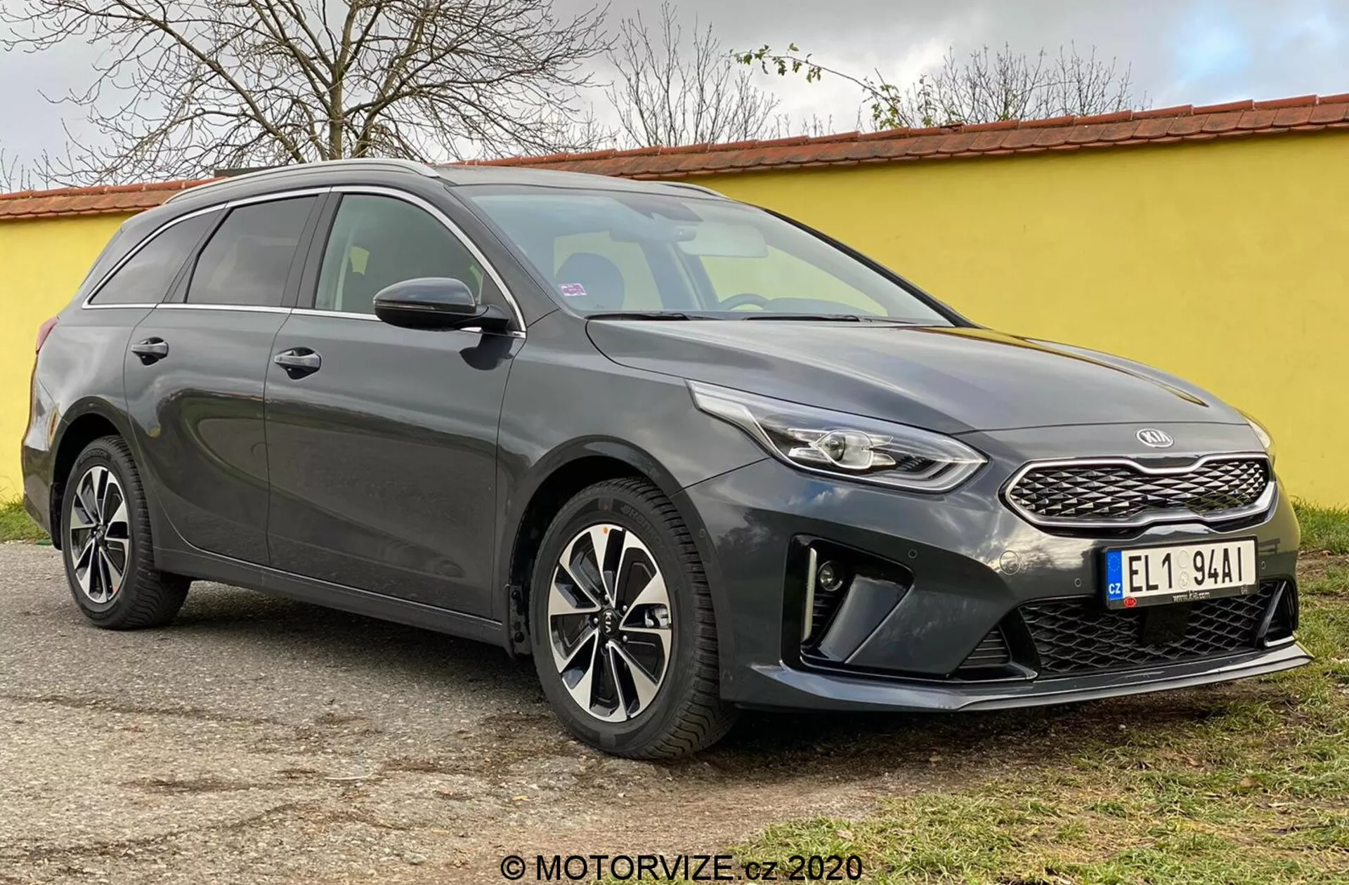 Obraz prezentuje Kia Ceed 2018, nowoczesne kombi uchwycone z przedniego kąta trzech czwartych, podkreślając jego przednią część, w tym strukturalny grill z charakterystycznym emblematem marki, wąskie, zaciągające się światła przednie z dziennymi światłami LED i przedni zderzak z obudowami świateł przeciwmgielnych i czujnikami parkowania. Eleganckie linie konturowe maski prowadzą do stromo pochylonej szyby przedniej, podkreślając aerodynamiczny profil samochodu. Widoczne są wieloramienne felgi aluminiowe i lusterka boczne z zintegrowanymi kierunkowskazami, wraz z opadającą linią dachu w kierunku tyłu i relingami dachowymi, podkreślając jednocześnie styl i funkcjonalność. Przyciemniane okna i linie nadwozia wzdłuż boku dodają rzeźbiarski wygląd. Samochód jest zaparkowany na płaskiej powierzchni na zewnątrz.