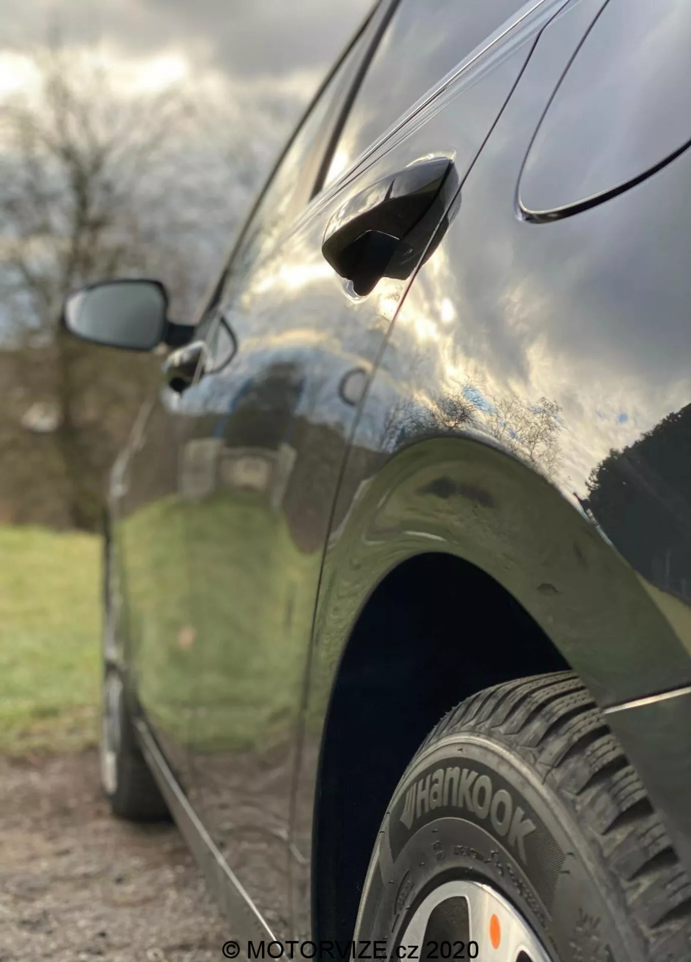 Zbliżenie boczne Kia Ceed 2018, ukazujące takie cechy jak tylne koło z oponą 'Hankook', aerodynamiczną klamkę drzwi i częściowo rozmyte boczne lusterko. Zdjęcie zrobione z niskiego kąta z małą głębią ostrości, co wyostrza tylne koło i dolną część drzwi, podczas gdy tło jest artystycznie rozmyte. Samochód ma ciemne, błyszczące wykończenie, które odbija niebo i otoczenie, ukazując eleganckie linie projektowe i dobrze utrzymaną karoserię.