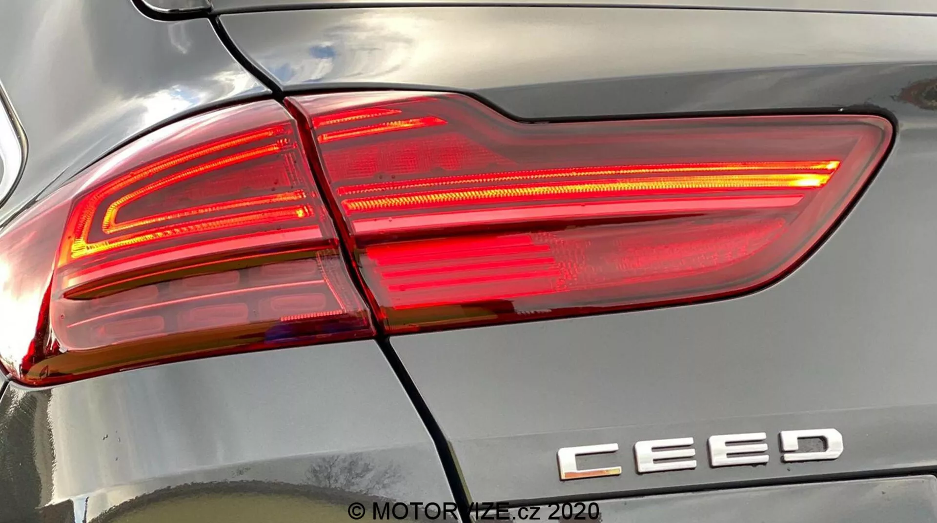 Zbliżenie szczegółowe tylnego prawego zespołu świateł w samochodzie Kia Ceed 2018, oświetlone, prezentujące nowoczesny design z ostrymi, kątowymi liniami, sekcjami soczewek czerwonych i przeźroczystych oraz odznaką modelu 'CEED'. Linia pozioma oddziela tylne światło od tylnego zderzaka. Odbicia na karoserii podkreślają krzywiznę powierzchni. Znak wodny 'MOTOR1.cz ©2020'.
