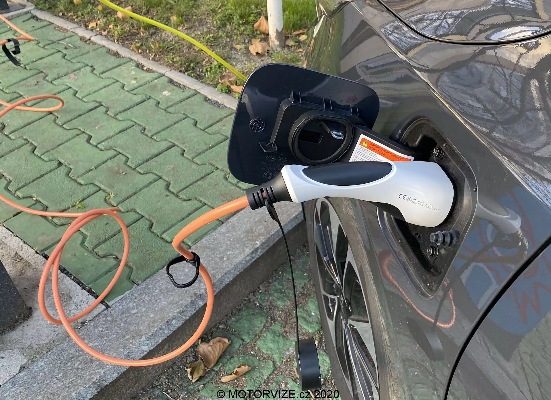 Zbliżony widok boczny szarego pojazdu elektrycznego (EV) podczas ładowania, uwydatniający otwarty port ładowania i podłączony pomarańczowy konektor EVSE. Nowoczesny biało-czarny konektor do ładowania posiada wskaźnik LED. Obraz skupia się na interakcji między samochodem a stacją ładowania, osadzony w zewnętrznym, utwardzonym obszarze z rozrzuconymi liśćmi.
