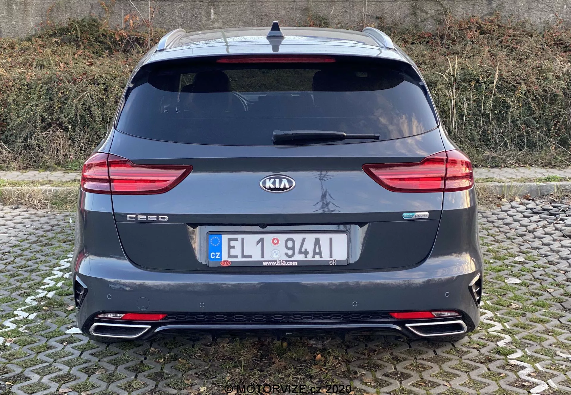 Widok tyłu Kia Ceed 2018 pokazujący całą tylną część z odznakami marki i modelu, nowoczesnymi tylnymi światłami LED, tylną szybą, podwójnymi końcówkami rur wydechowych, tylnym zderzakiem z odblaskami, spojlerem na dachu, anteną i tylnymi czujnikami parkowania. Obraz jest wyraźny, wykonany na wysokości oczu przy naturalnym oświetleniu w pochmurnych warunkach.