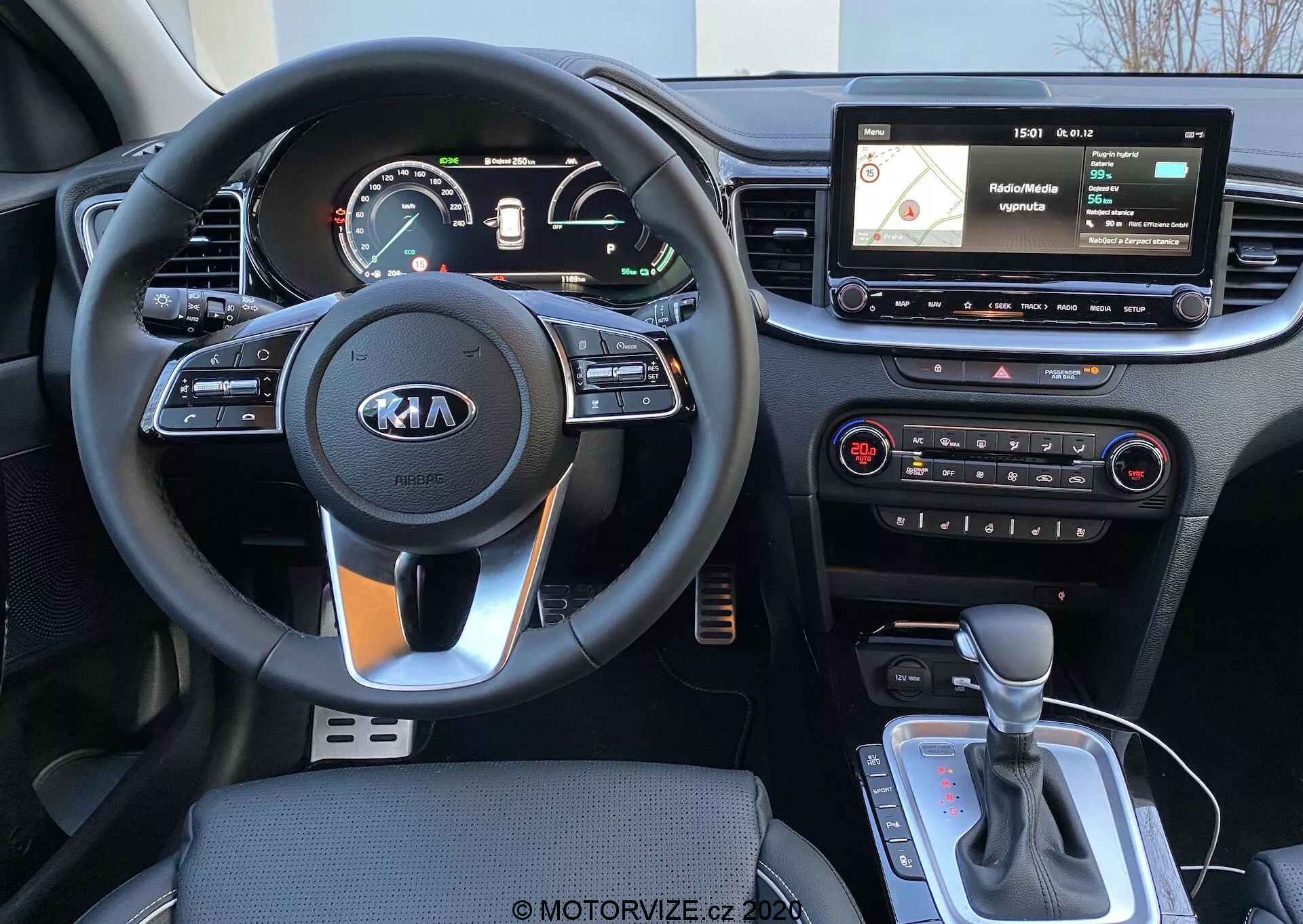 Widok wnętrza Kia Ceed 2018 z perspektywy kierowcy, przedstawiający trójramienną kierownicę z logo Kia, przyciski na kierownicy, analogowo-cyfrowy zestaw wskaźników, unoszący się ekran infotainment z nawigacją i interfejsem medialnym, fizyczne przyciski klimatyzacji oraz dźwignię automatycznej skrzyni biegów z możliwym selektorem trybu jazdy lub elektrycznym hamulcem ręcznym.