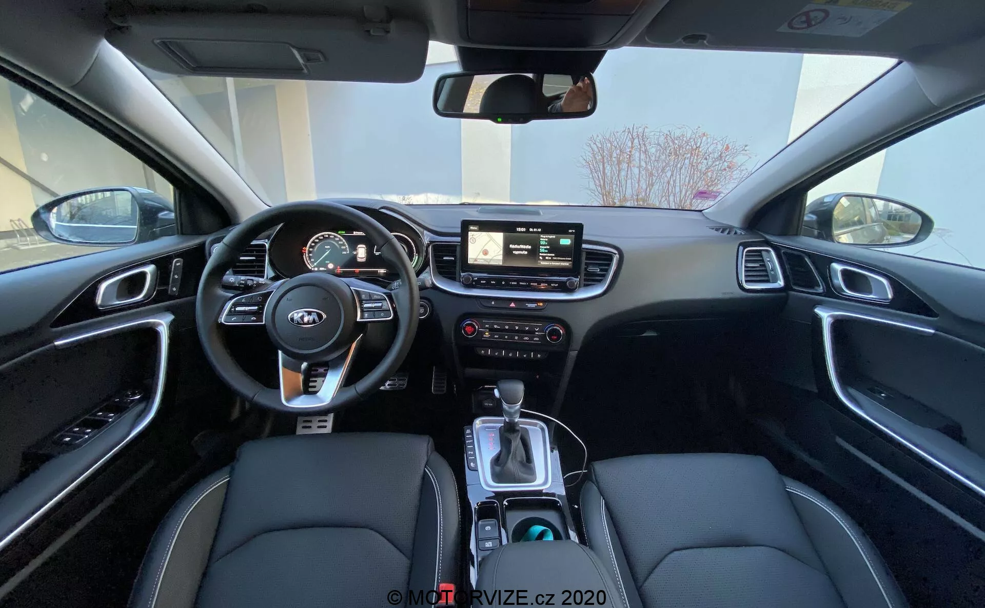 Szerokokątny widok wnętrza Kia Ceed 2018 z tylniego siedzenia, ukazujący obszar kierowcy, w tym kierownicę z zamontowanymi kontrolkami, zestaw wskaźników z cyfrowymi i analogowymi miernikami, centralny system infotainment z ekranem dotykowym, ręczne kontrole dla systemu infotainment i klimatyzacji, oraz konsola centralna z dźwignią zmiany biegów, selektorem trybu jazdy, uchwytami na kubki i czarnym wnętrzem, posiadającym fotele z syntetycznej skóry z białymi przeszyciami oraz wykończeniami z aluminium szczotkowanego.
