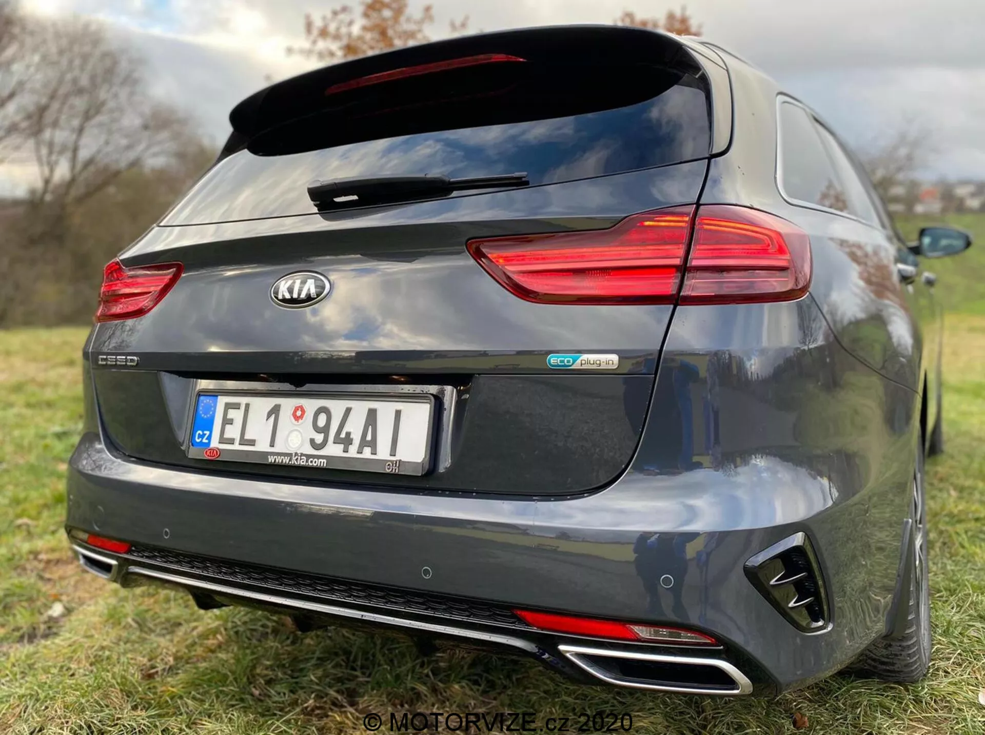 Tylny widok ciemnoszarego modelu Kia Ceed Proceed 2018, z detalami takimi jak poziomo zorientowane tylne światła LED, tylni spoiler zamontowany na dachu, podwójne chromowane końcówki wydechowe, tylny zderzak w stylu dyfuzora i centralne emblematy w tym 'Kia' i 'GT Line'. Kamera cofania nad centralnie umieszczoną tablicą rejestracyjną sugeruje asystę parkowania, a płytka głębia ostrości podkreśla markę i identyfikatory modelu samochodu na tle trawiastym.