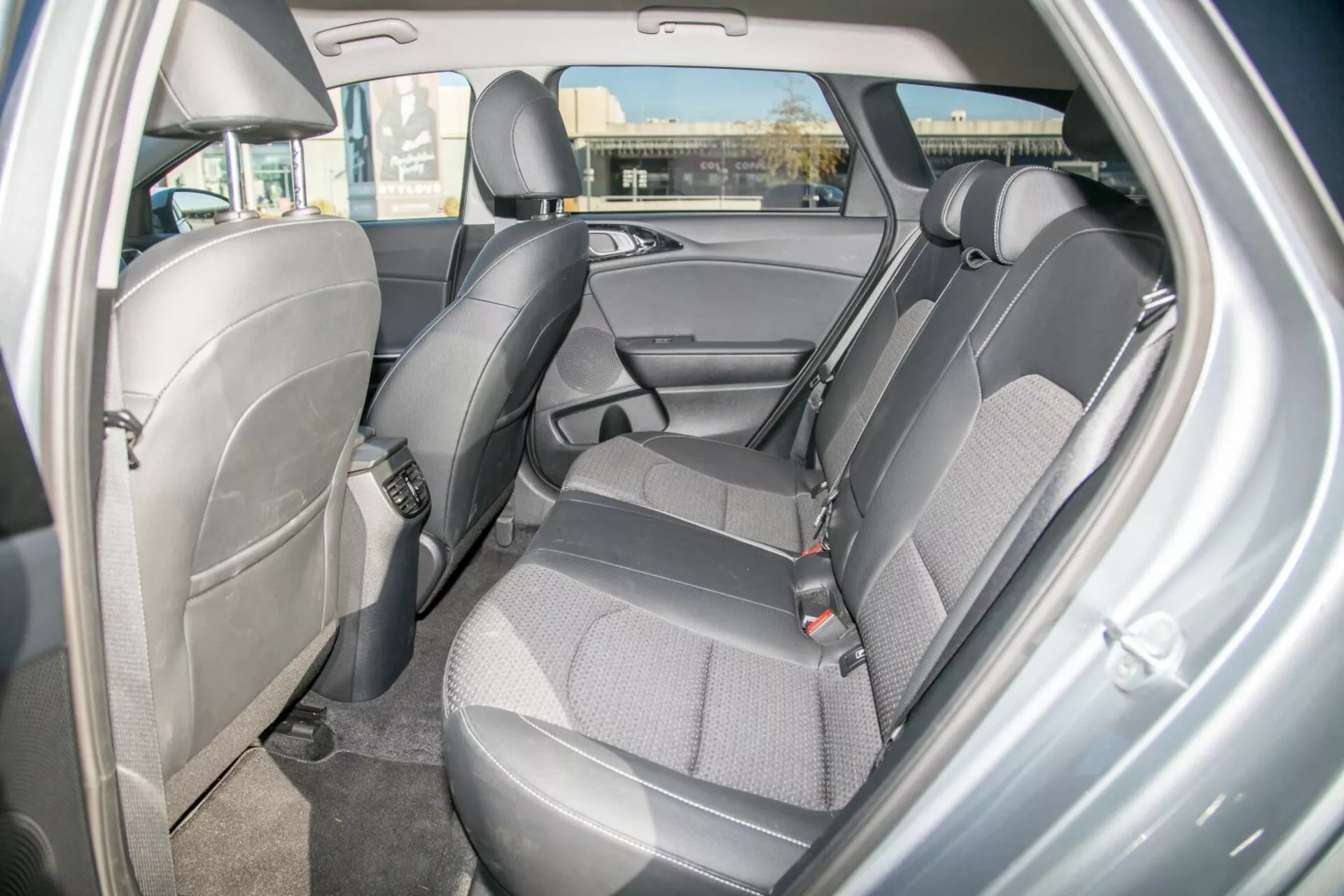 Widok wnętrza Kia Cee'd 2009 prezentujący design i funkcje tylnej części siedzenia, podkreślając wytrzymałą tapicerkę z tkaniny w dwóch odcieniach, widoczne pasy bezpieczeństwa, regulowane zagłówki, panel drzwi z miejscem do przechowywania, potencjalne kotwy do fotelika dziecięcego, wykładzinę podłogową i ambientowe oświetlenie kopułkowe z perspektywy tylnych drzwi po stronie prawej.