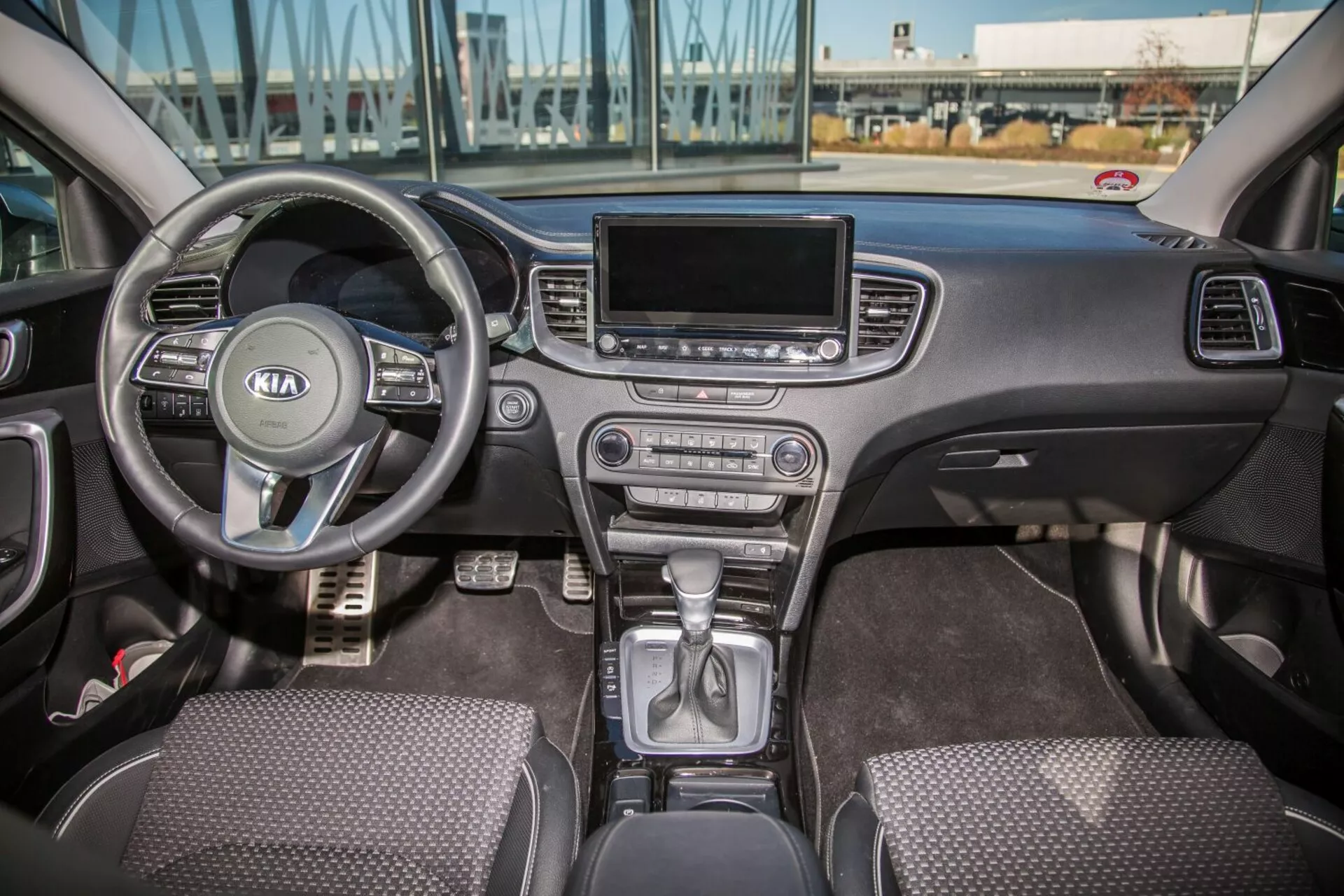 Wnętrze Kia Cee'd z 2009 roku prezentujące pełną przednią kabinę w tym wielofunkcyjną kierownicę z logiem Kia, część zespołu wskaźników za kierownicą, dźwignię zmiany biegów w pozycji park na konsoli środkowej, przyciski kontroli klimatyzacji, ekran systemu infotainment, siedzenia pokryte tkaniną, oraz kontrolery drzwi kierowcy i pasażera w naturalnym świetle dziennym