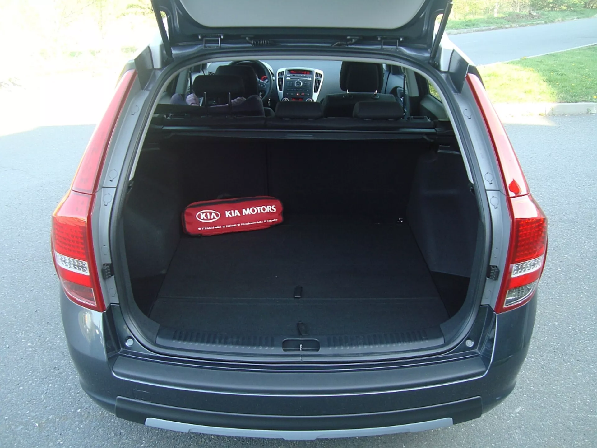 Tylna część bagażnika hatchbacka Kia Cee'd z 2009 roku z otwartą klapą ukazującą przestronny, pusty obszar ładunkowy, chowaną osłonę, wewnętrzne wykładziny tekstylne i charakterystyczne logo Kia Motors na czerwonej osłonie ochronnej, prezentujące cechy projektowe, bezpieczeństwa i technologii pojazdu.