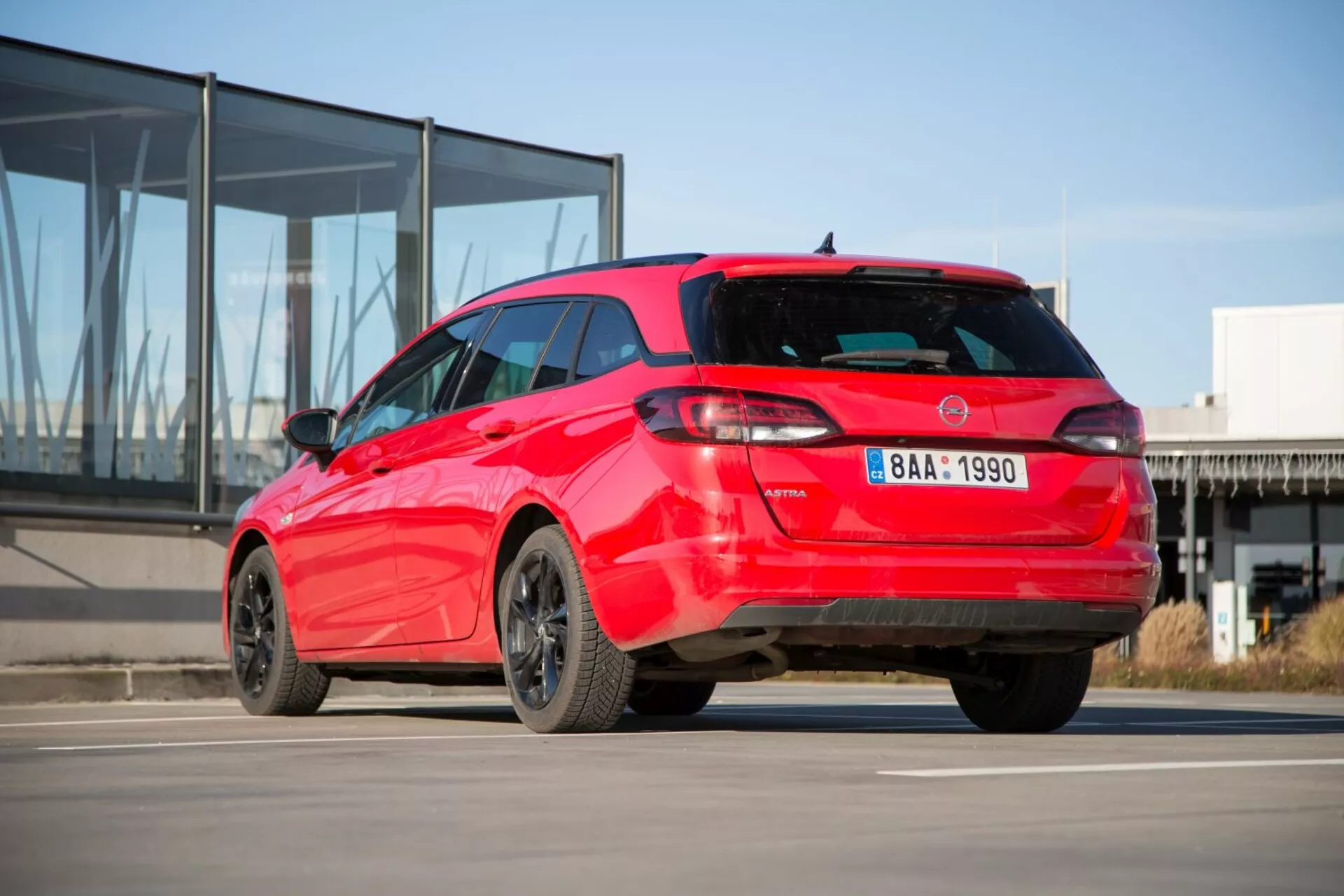 Czerwony hatchback Kia Ceed 2009 wyświetla swój sportowy widok z tyłu trzy czwarte na betonie z tłem nowoczesnego budynku ze szklaną fasadą, pokazując wyróżniający się wzór tylnych świateł, spoiler dachowy, antenę w kształcie płetwy rekina, felgi aluminiowe i możliwe czujniki parkowania.