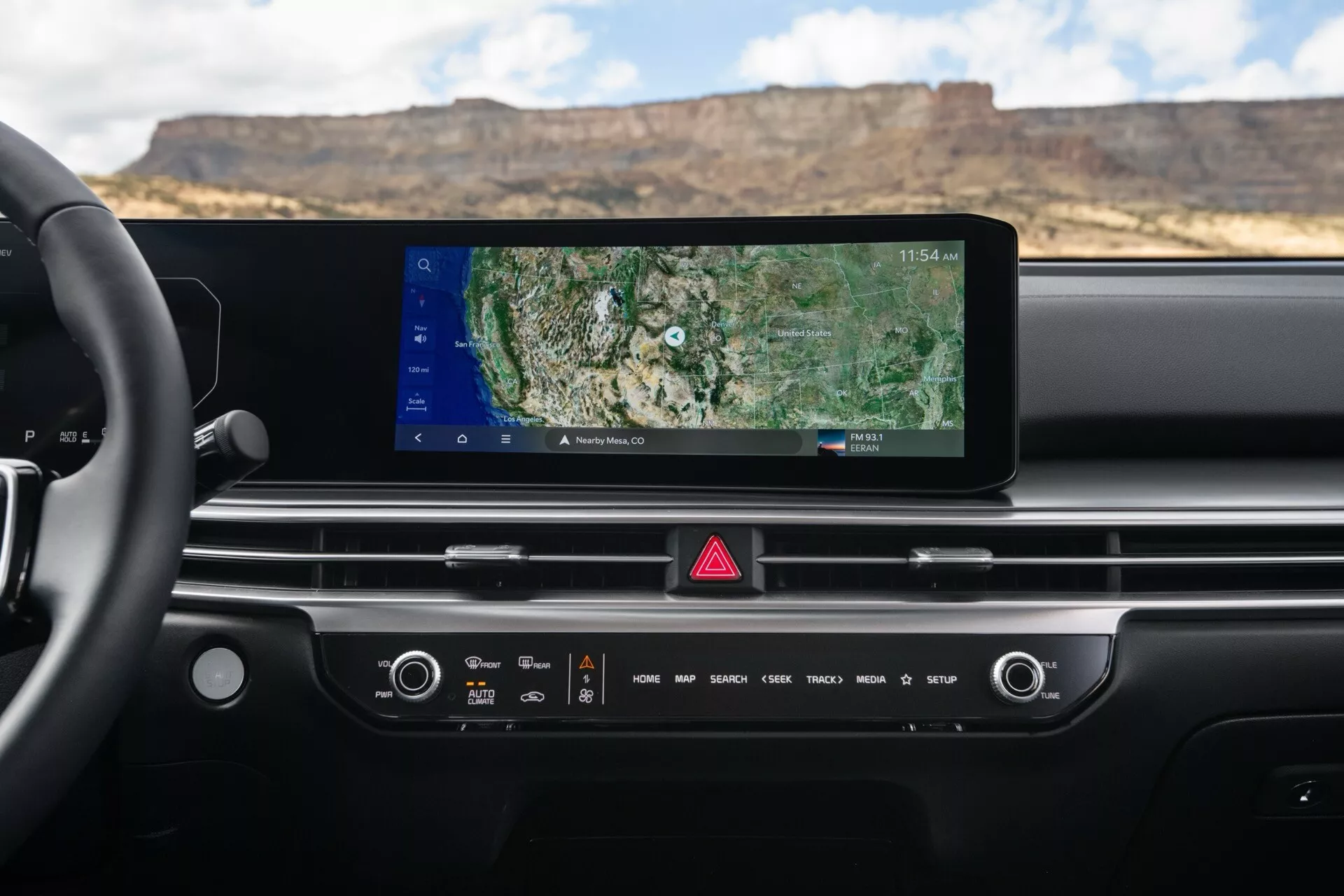 Wnętrze deski rozdzielczej Kia Sorento MQ4 facelift 2024 z szerokim ekranem dotykowym systemu infotainment z nawigacją GPS, ręcznymi i dotykowymi elementami sterującymi klimatyzacją i dźwiękiem, przyciskiem świateł awaryjnych i minimalistycznym designem z zintegrowanymi nawiewami powietrza, widziane z perspektywy kierowcy