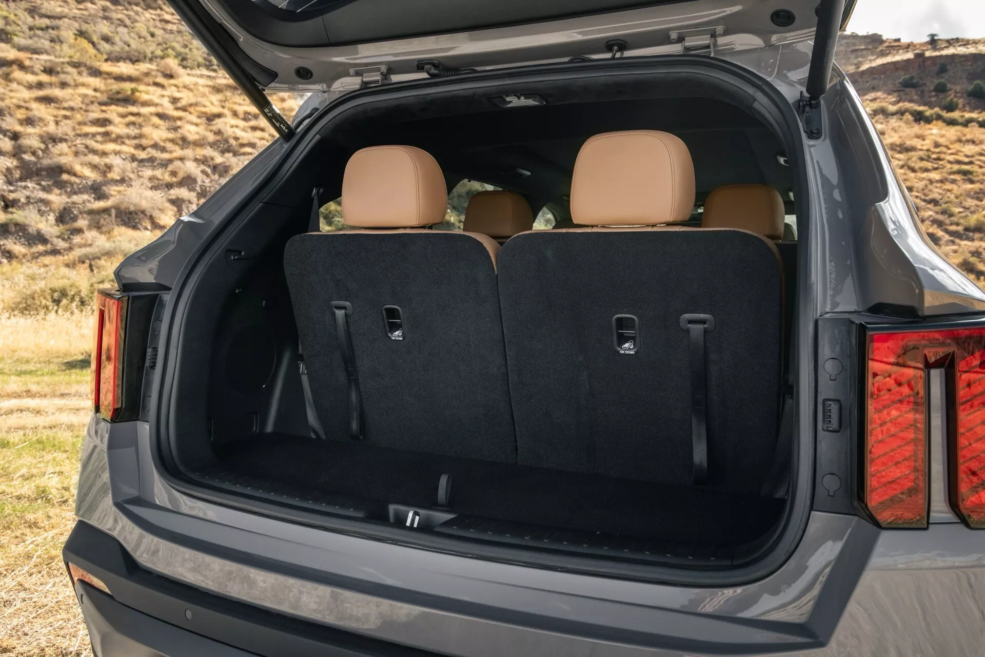 Facelifting Kia Sorento MQ4 2024 prezentuje przestronny bagażnik z otwartym tylnym hatchbackiem, tylne siedzenia dzielone w stosunku 60/40, ergonomiczną płaską podłogę ładowania, jasne wykończenie wnętrza i wygodne boczne kieszenie, na tle naturalnego krajobrazu.