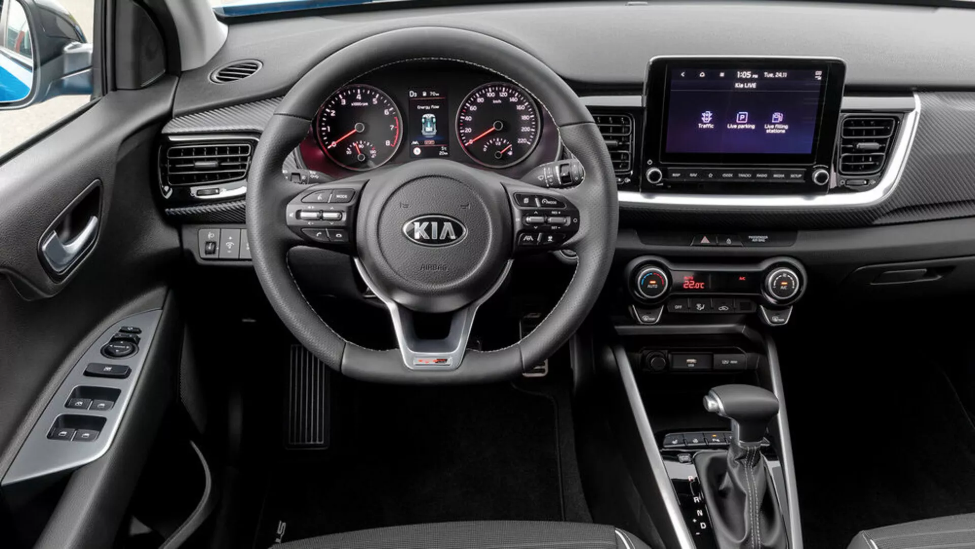 Widok wnętrza Kia Stonic z 2017 roku z perspektywy pasażera, przedstawiający kierownicę z trzema ramionami i zamontowanymi sterowaniami, analogowo-cyfrowy zestaw wskaźników, system infotainment z ekranem dotykowym, kontrolę klimatyzacji, automatyczną skrzynię biegów oraz selektor trybu jazdy.
