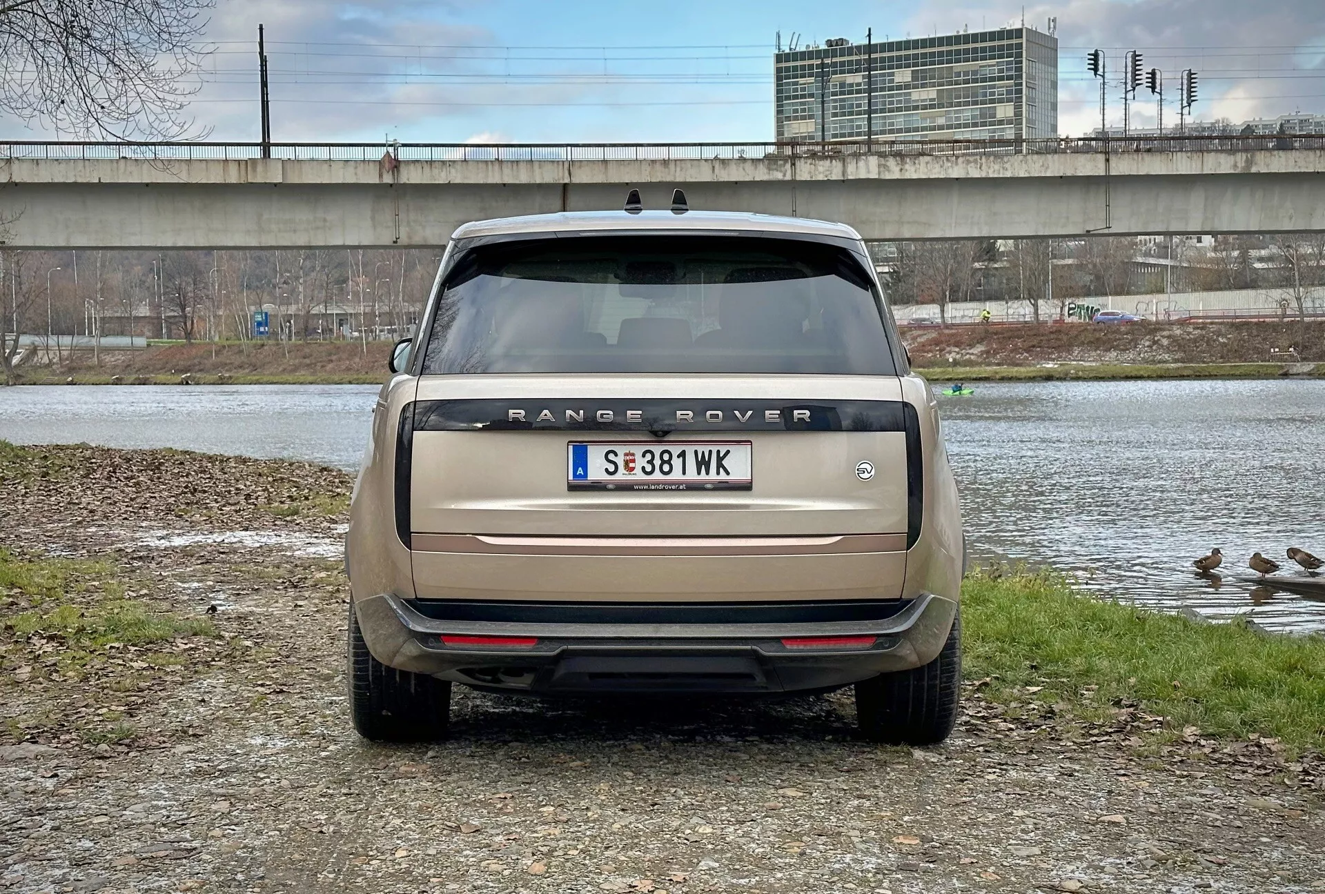 Tylny widok Range Rovera 2021 z bezpośrednim kątem patrzenia na wysokości oczu, zaparkowanego w pobliżu zbiornika wodnego z kaczkami i wiaduktem w tle, przedstawiający nazwę modelu, tylne światła, czujniki parkowania oraz podwójny układ wydechowy w matowym beżu