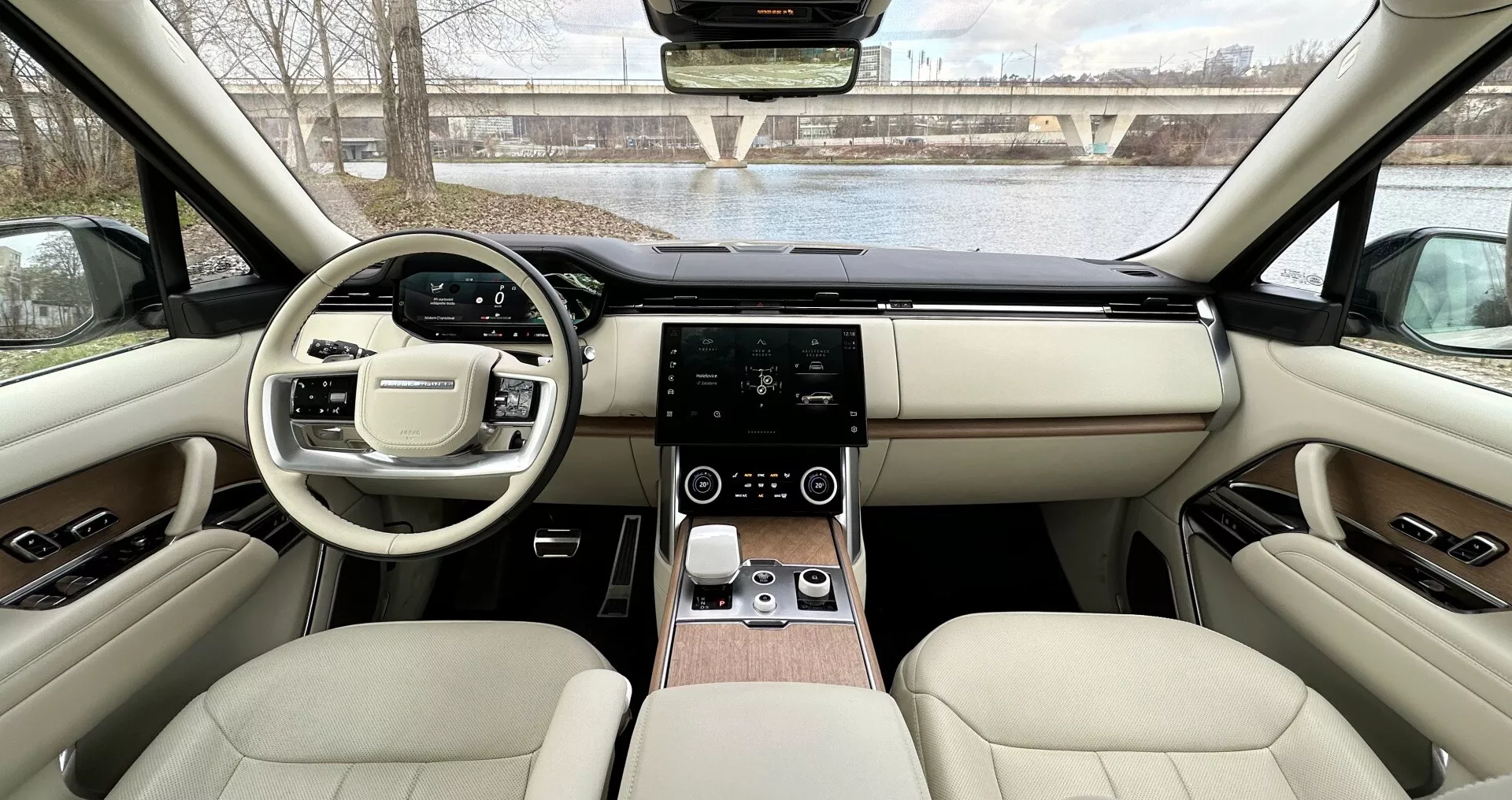 Widok wnętrza Land Rover Range Rover 2021 z tylnych siedzeń w kierunku deski rozdzielczej pokazujący symetryczny design, kierownicę z przyciskami wielofunkcyjnymi, dotykowy system infotainment, cyfrowe sterowanie klimatyzacją, luksusową tapicerkę skórzaną z akcentami drewna i zaawansowane funkcje technologiczne