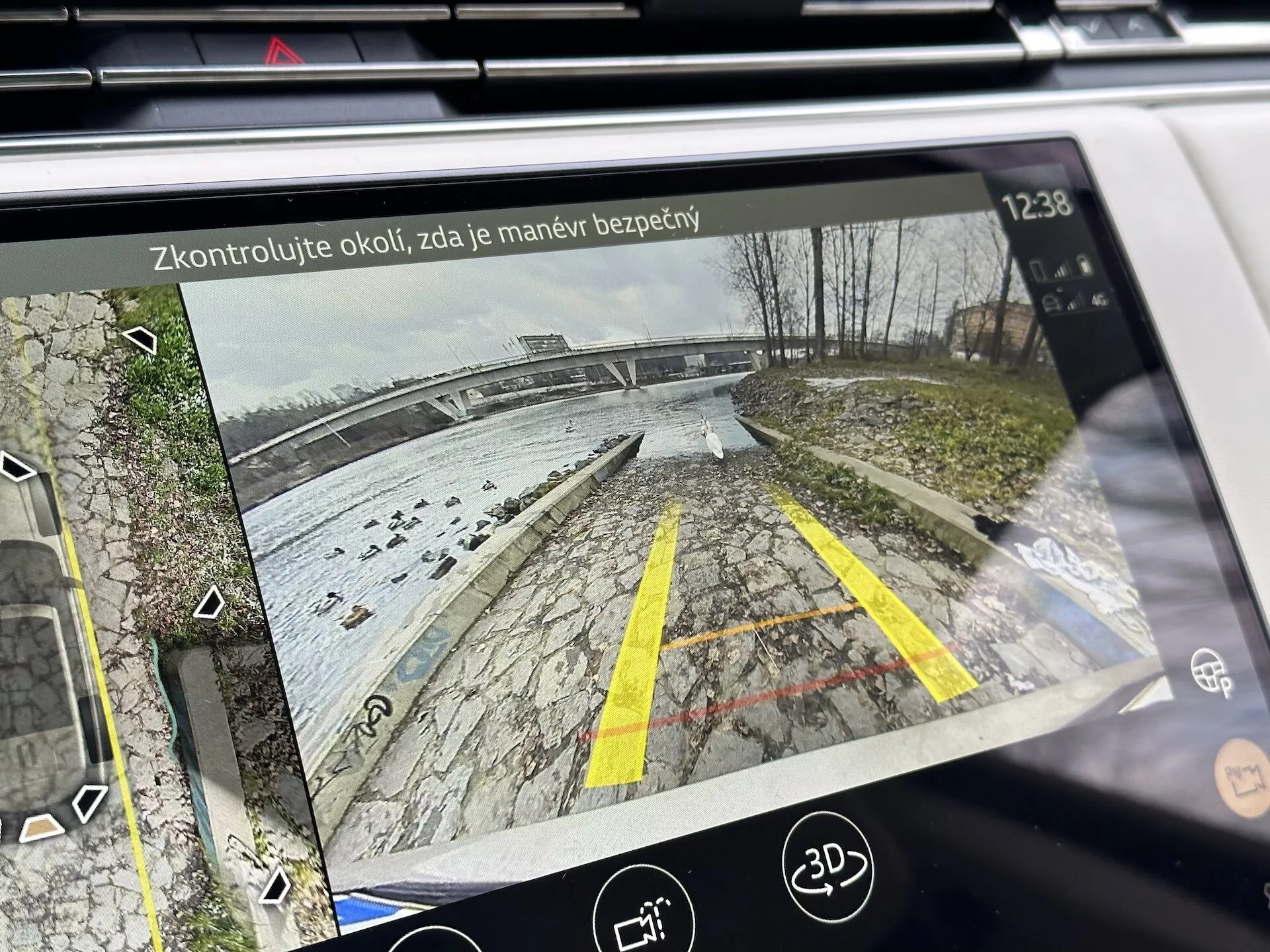 Wyświetlacz systemu infotainment Land Rover Range Rover 2021 pokazujący tylny widok z kamery cofania z dynamicznymi liniami prowadzącymi na parkingu w pobliżu wody, szczegółowo z roślinnością, ikonami wsparcia przy parkowaniu i tekstem przypominającym o bezpieczeństwie