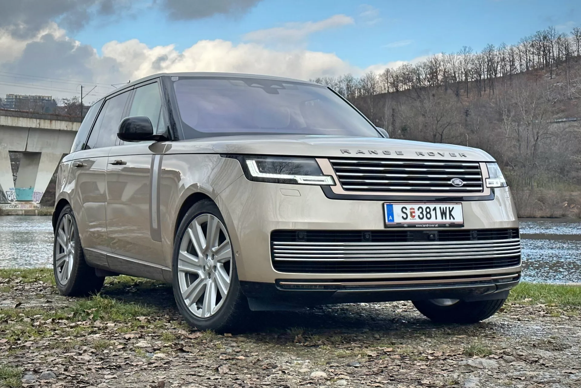 Luksusowy SUV Land Rover Range Rover 2021, widok z przodu i trzech czwartych ukazujący chromowaną atrapę, reflektory LED, felgi aluminiowe o wielu ramionach i aerodynamiczny design, zaparkowany na zewnątrz blisko rzeki i mostu