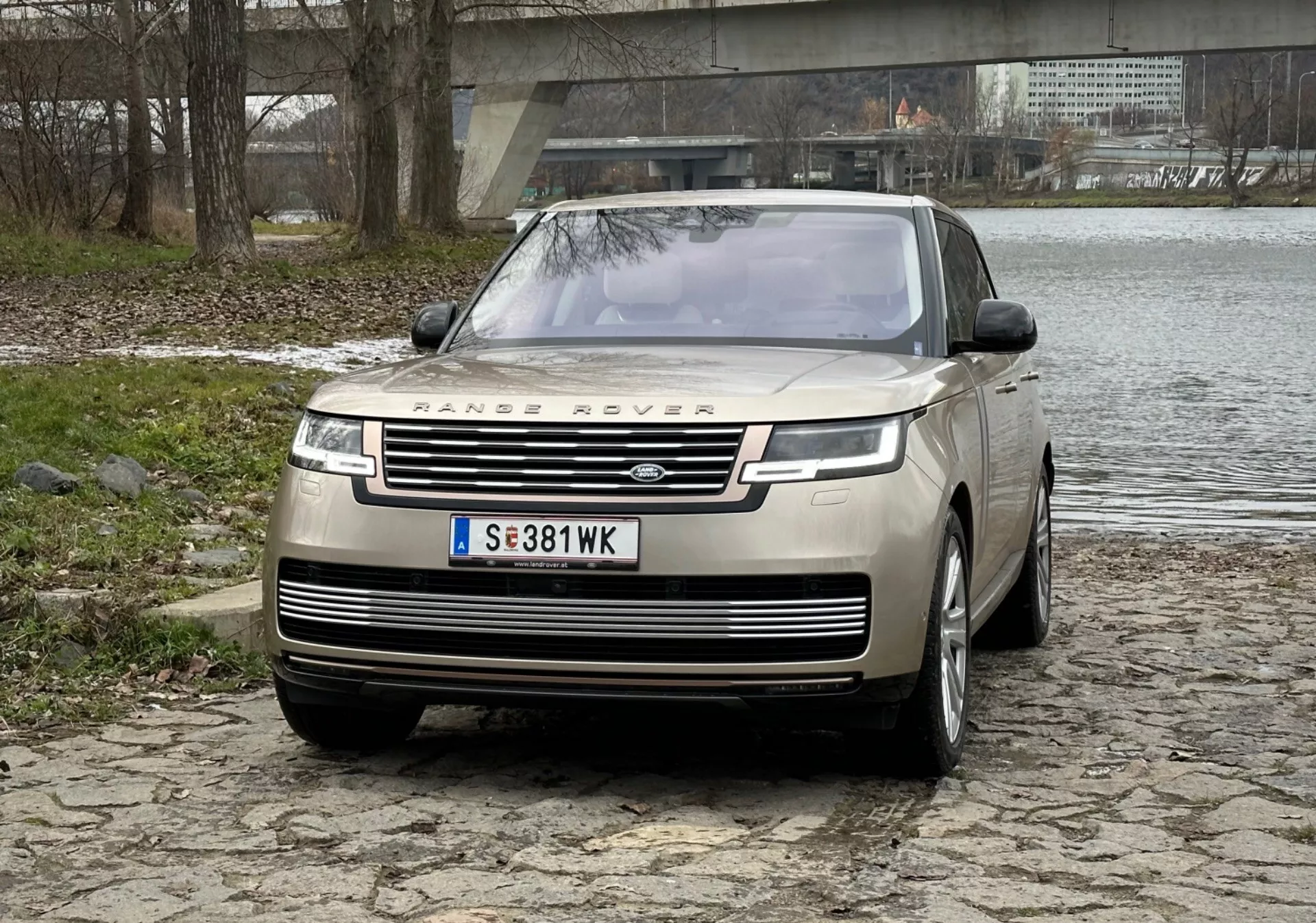 Luksusowy SUV Range Rover 2021 w kolorze srebrnym, wyeksponowana przednia atrapa z brandingiem, eleganckie światła do jazdy dziennej LED, przedni zderzak z czujnikami parkowania i design maski typu clamshell na brukowanej powierzchni w pobliżu rzeki