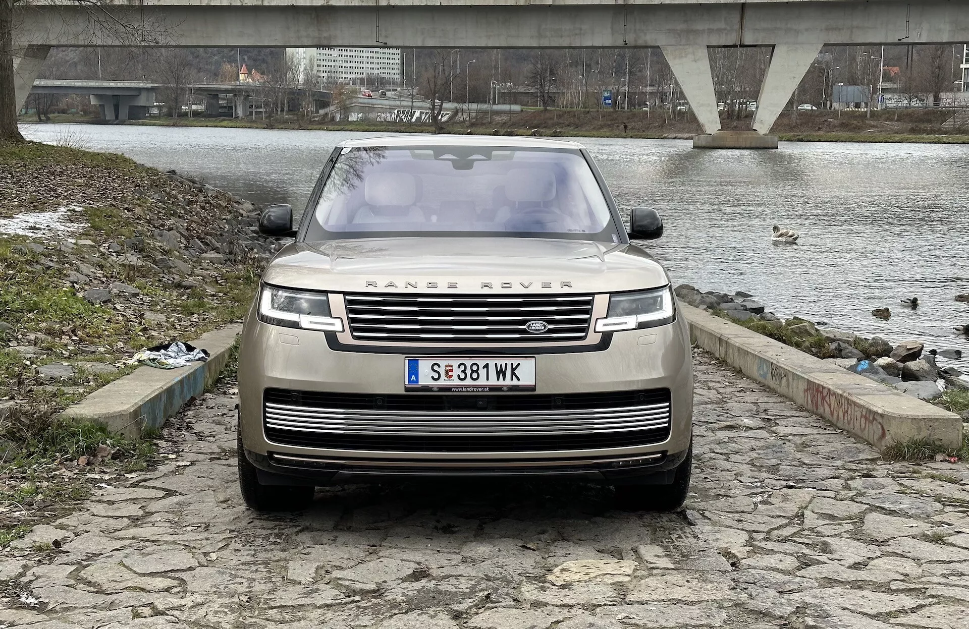 Land Rover Range Rover 2021 ukazany z przodu, z lekko niskiego kąta, podkreślający grill, przedni zderzak i maskę, z ikonicznym emblematem RANGE ROVER, nowoczesnymi światłami LED i zaawansowaną technologią asystenta kierowcy, na tle rzeki lub jeziora, mostu i pochmurnego nieba