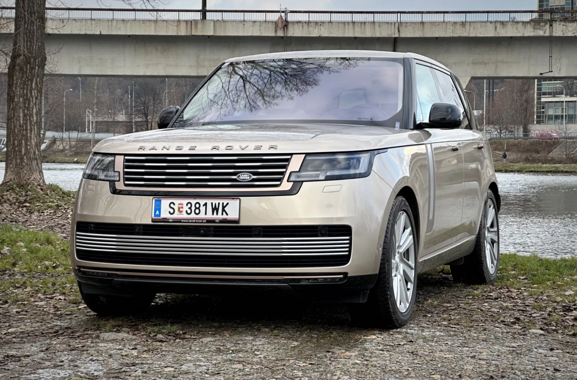Range Rover 2021 widziany z imponującej perspektywy trzy czwarte z przodu, pokazujący charakterystyczną kratkę, eleganckie reflektory LED i solidną płytę ochronną z przodu na tle miejskiego parku, podkreślając luksus i zdolności terenowe.