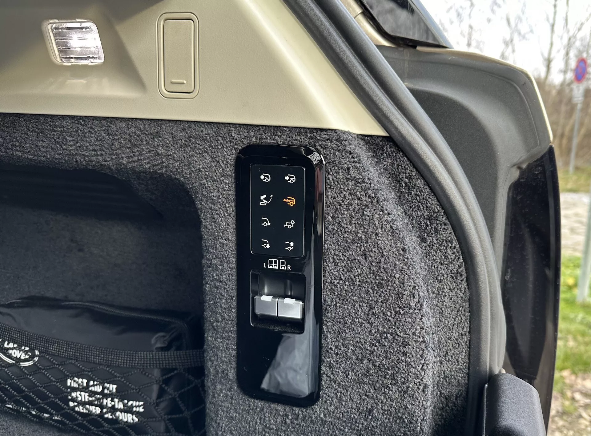 Zbliżenie na elektroniczny panel sterowania bagażnika w interiorze Land Rover Range Rover 2021, pokazujący przyciski do operacji takich jak elektroniczne otwieranie i zamykanie, kontrola pokrywy ładunku, potencjalnie światło bagażnika lub gniazdo zasilania, regulacje tylnych siedzeń z bagażnika i awaryjne zwolnienie, umieszczone przeciwko wykładzinie bocznej bagażnika i jasnego sufitu z oprawą świetlną ładunku.