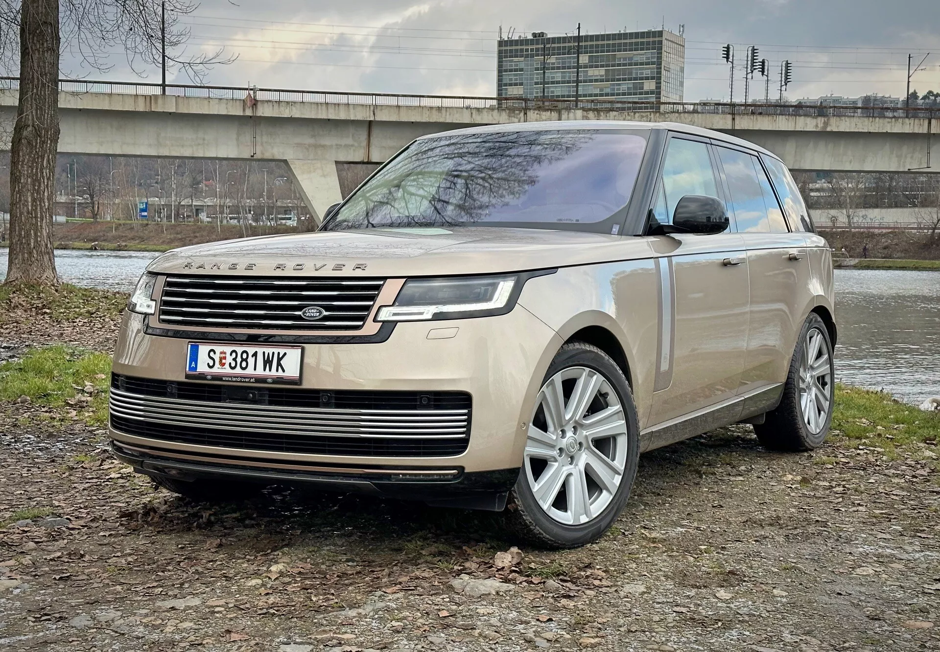 Luksusowy SUV Land Rover Range Rover 2021 zaparkowany na mieszanej nawierzchni ziemi i trawy z betonową konstrukcją i roślinnością w tle, prezentujący widok z przodu podkreślający reflektory LED, koła z stopu i nowoczesne funkcje aerodynamiczne